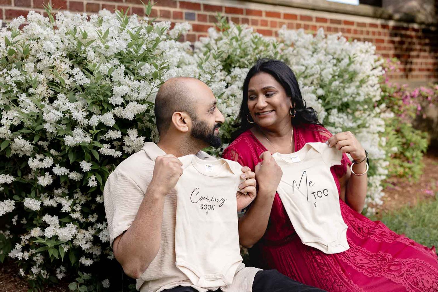 Couple sitting together by flowering bushes showing matching white coffee mugs in a garden setting.