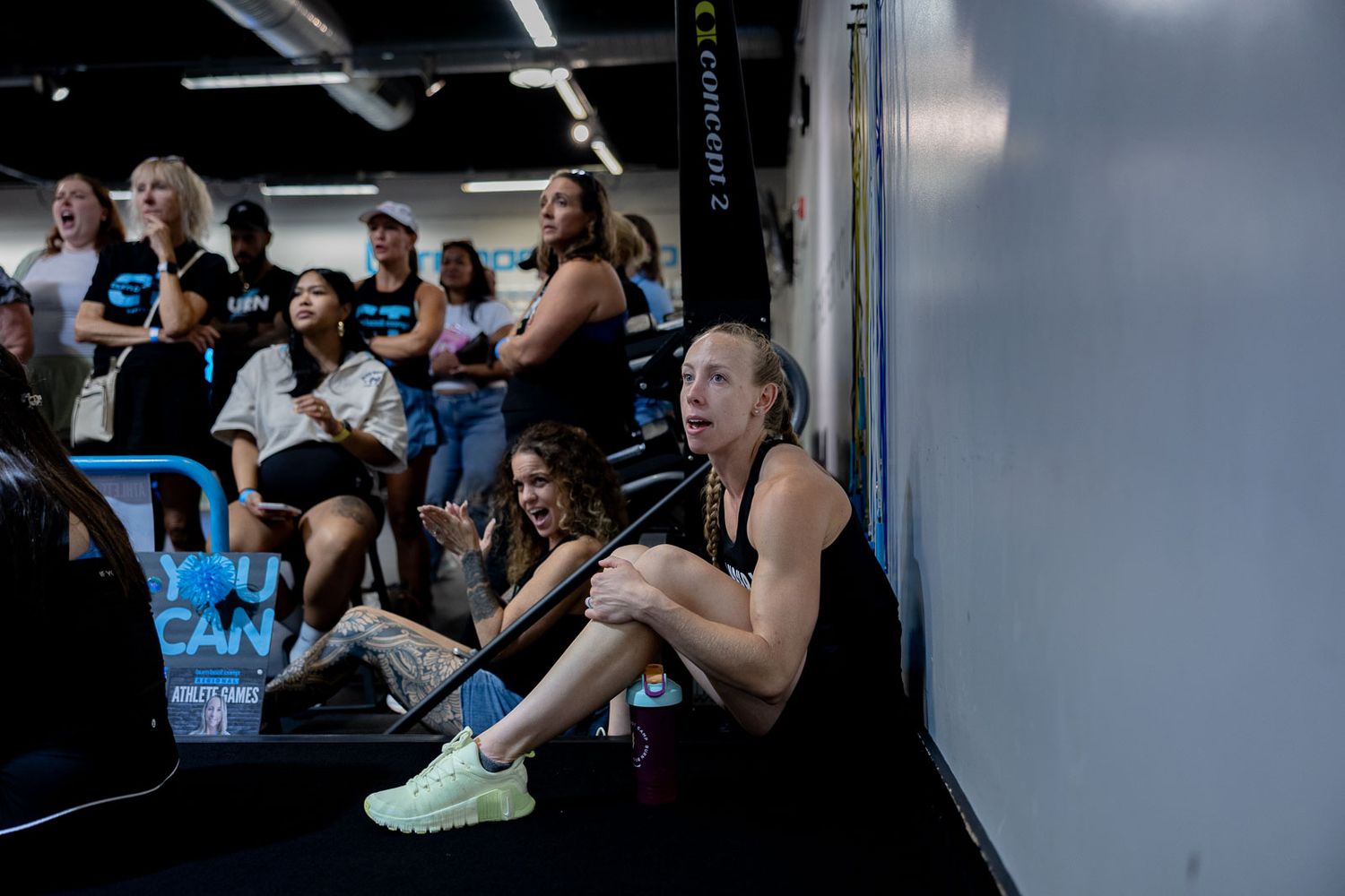 Athletes resting and watching during an intense workout session at a CrossFit style gym with gym equipment visible.