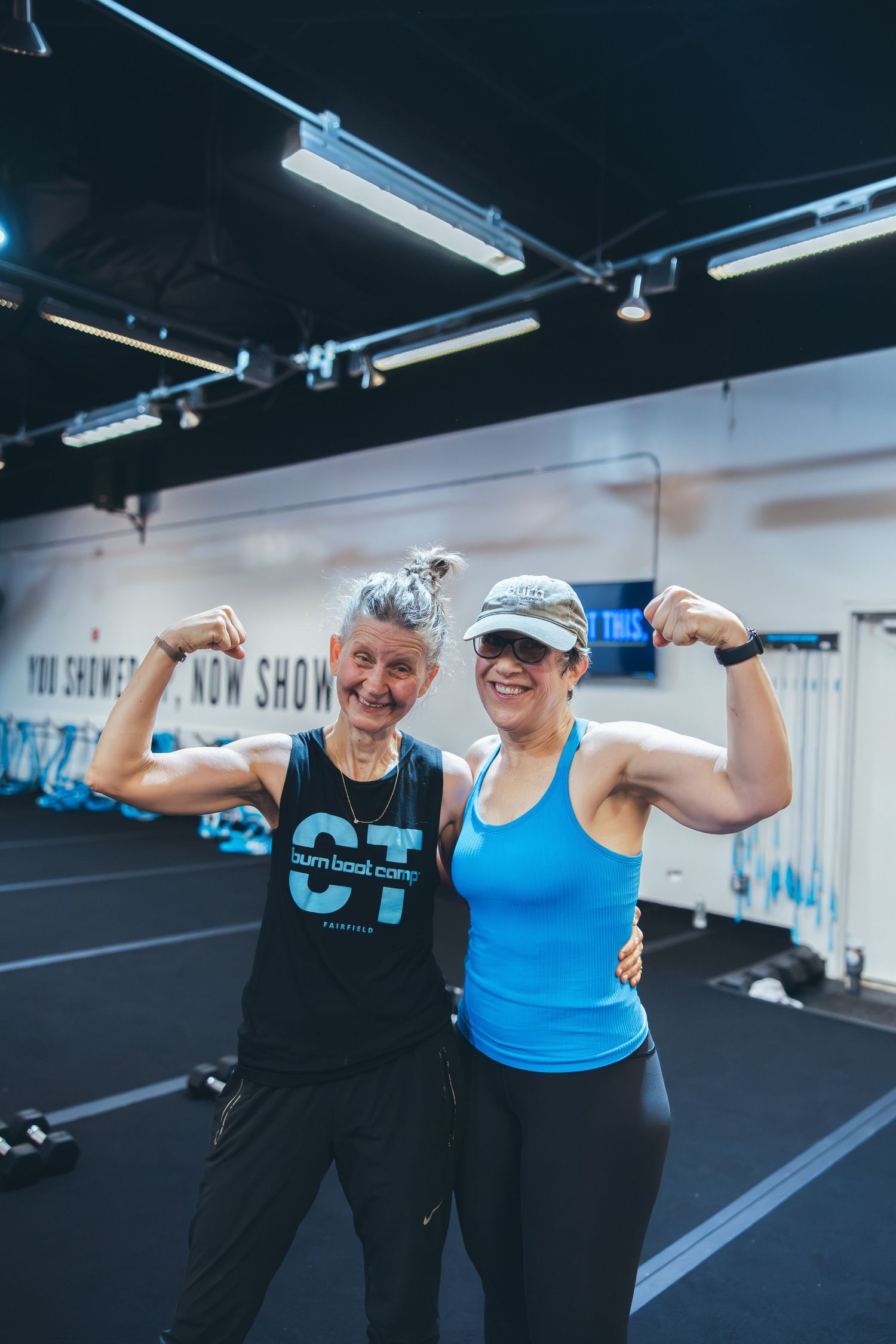 Two athletic individuals flexing their biceps and smiling at a fitness center.