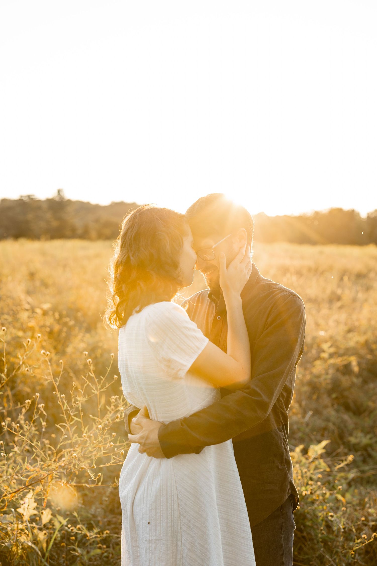 A romantic couple embraces in a golden sunlit field during sunset, creating a dreamy silhouette against the warm light.