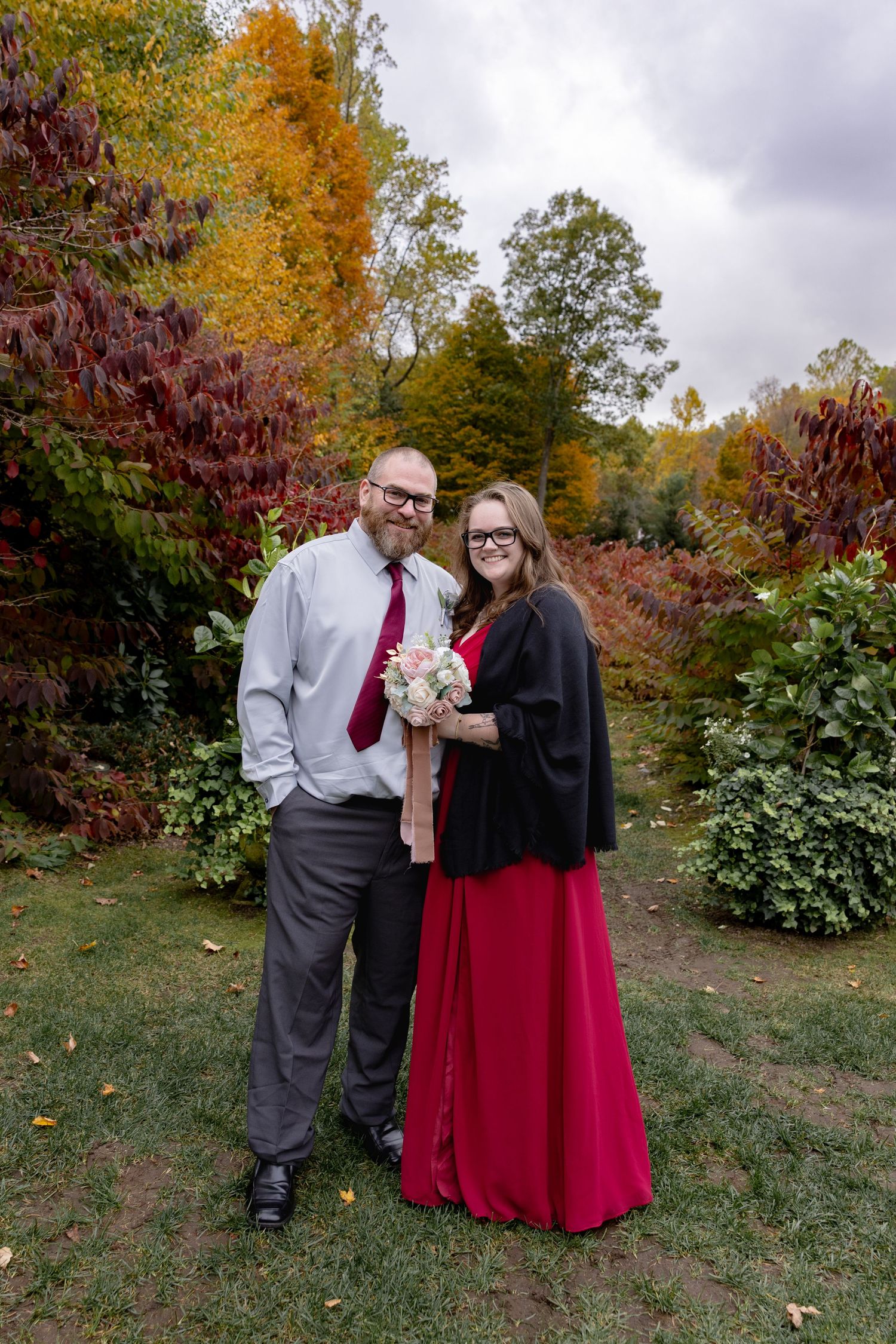 A couple stands together on an autumn day surrounded by colorful fall foliage while holding a small bouquet.