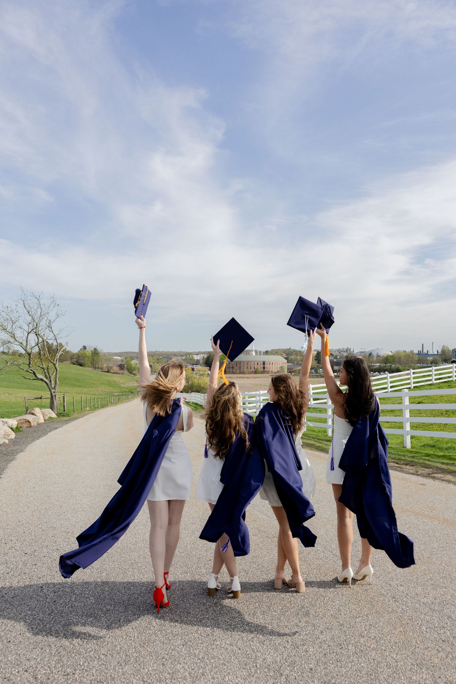 Four graduates in navy blue caps and gowns celebrate on a rural road by raising their graduation caps against a blue sky.