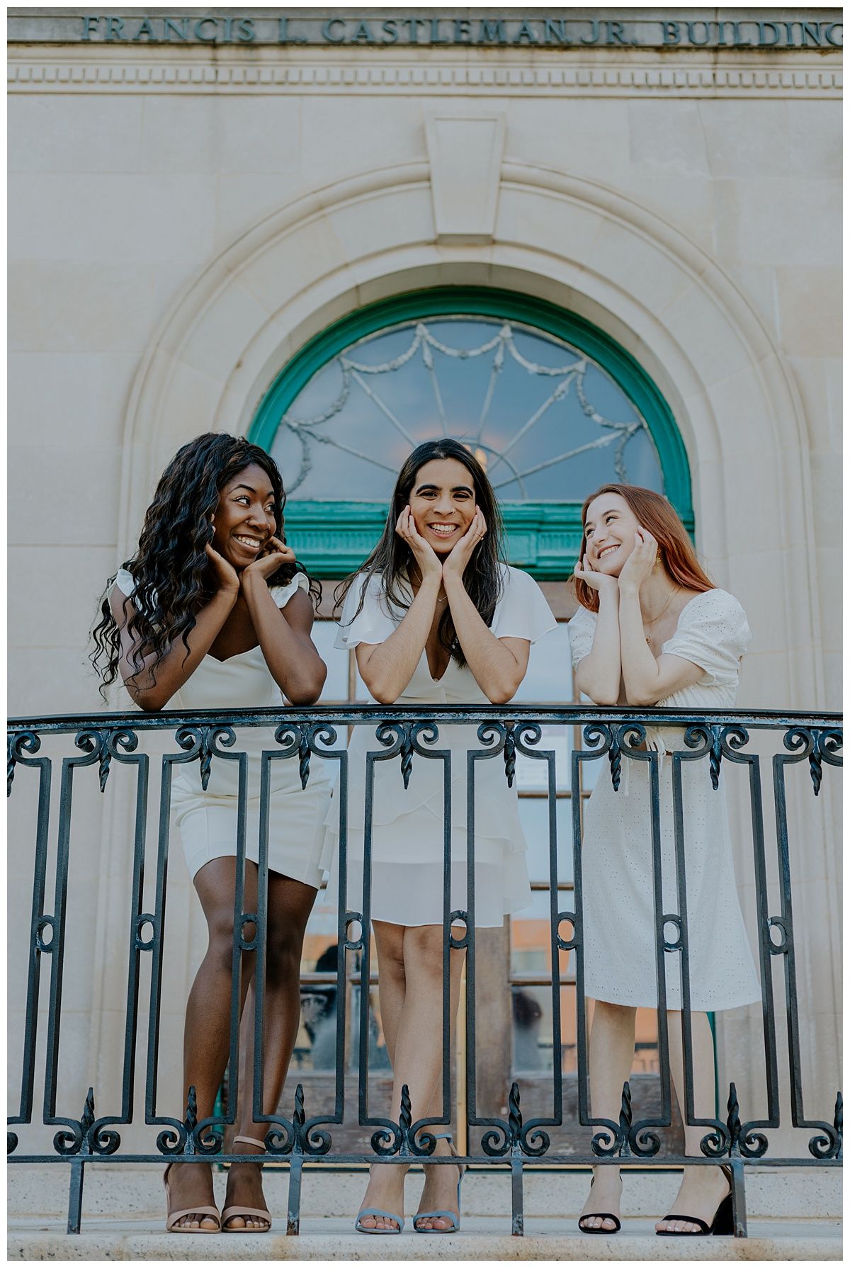 Three women in white outfits share joyful expressions on a balcony with ornate ironwork and arched doorway.