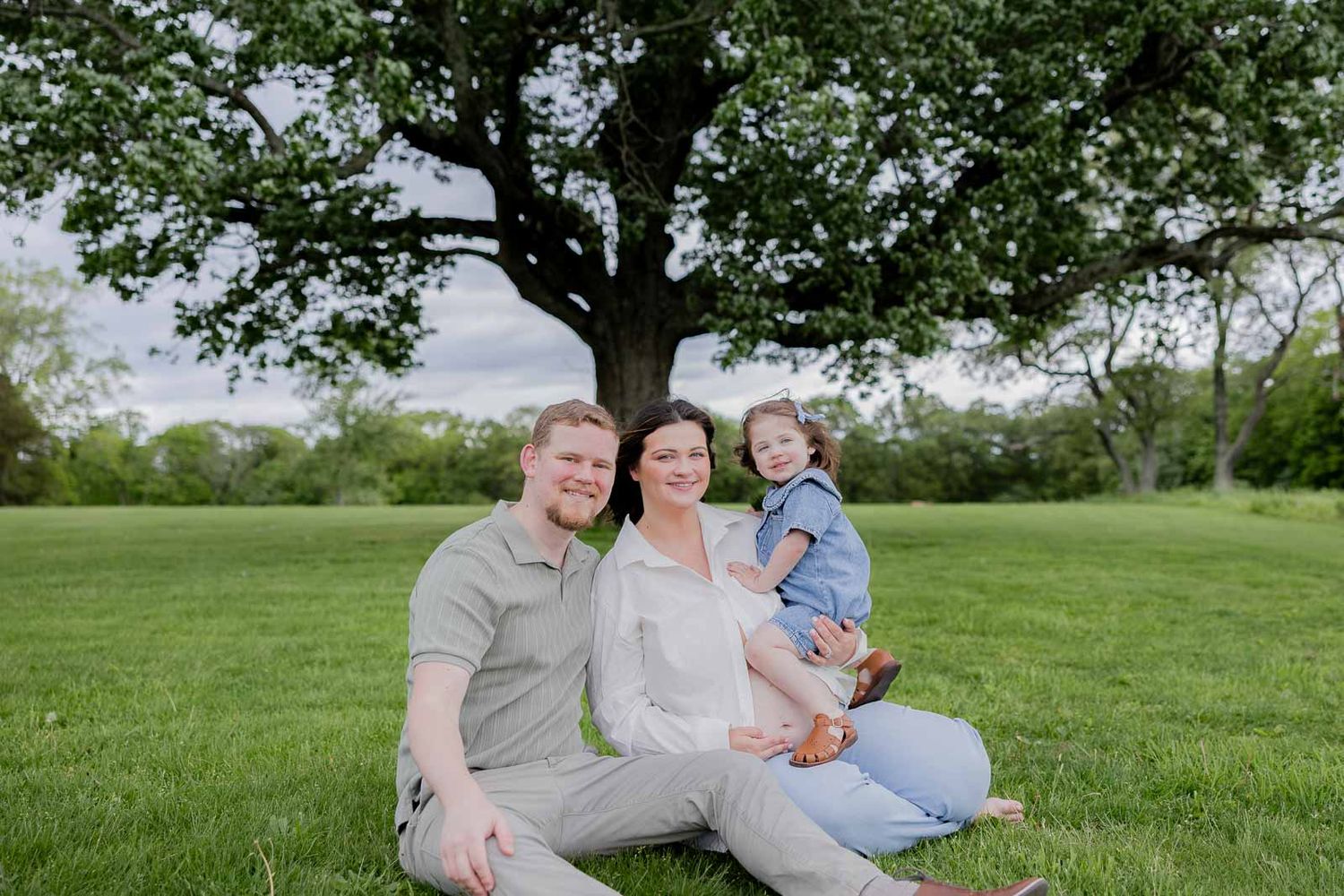 A family sits together on grass under a large tree during a spring outdoor photo session.