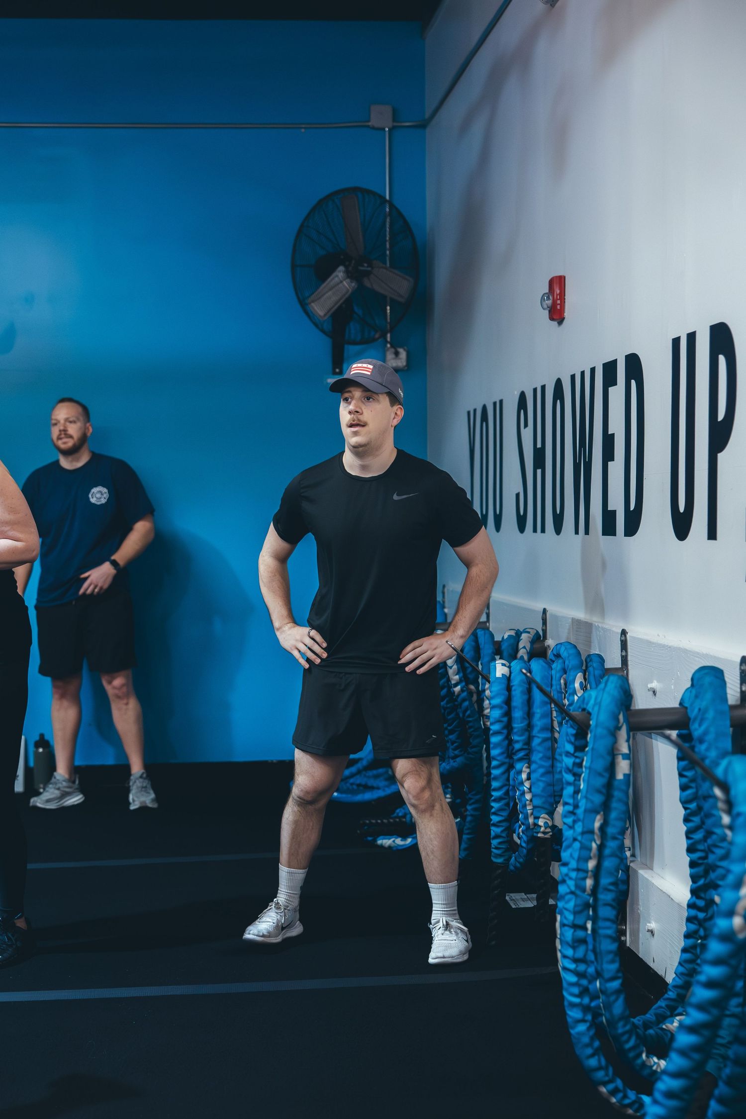 Blue and white gym interior with exercise ropes mounted on wall and large industrial fan.
