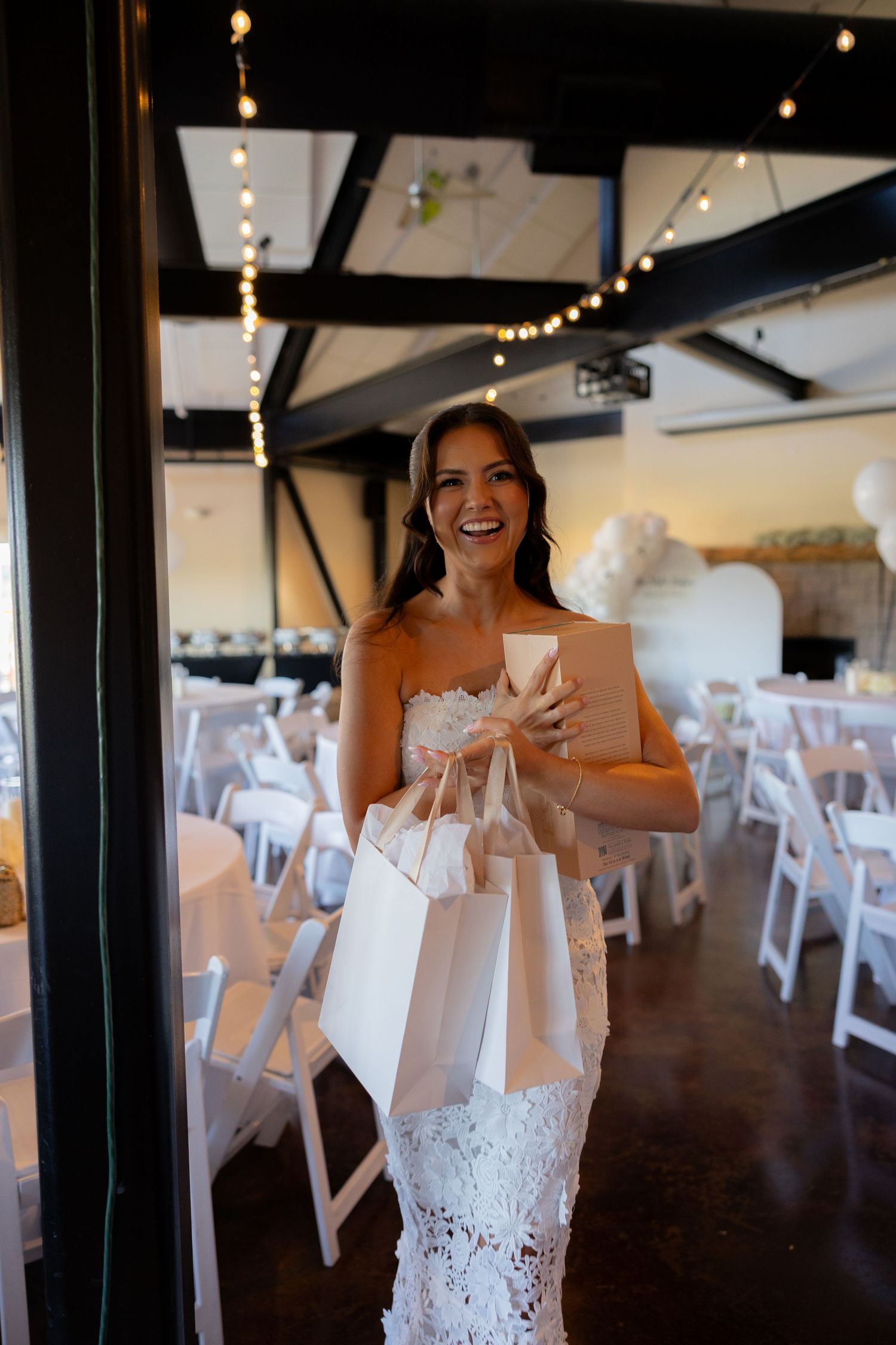 A bride in a white lace dress smiles while holding white gift bags in an indoor venue with white chairs.