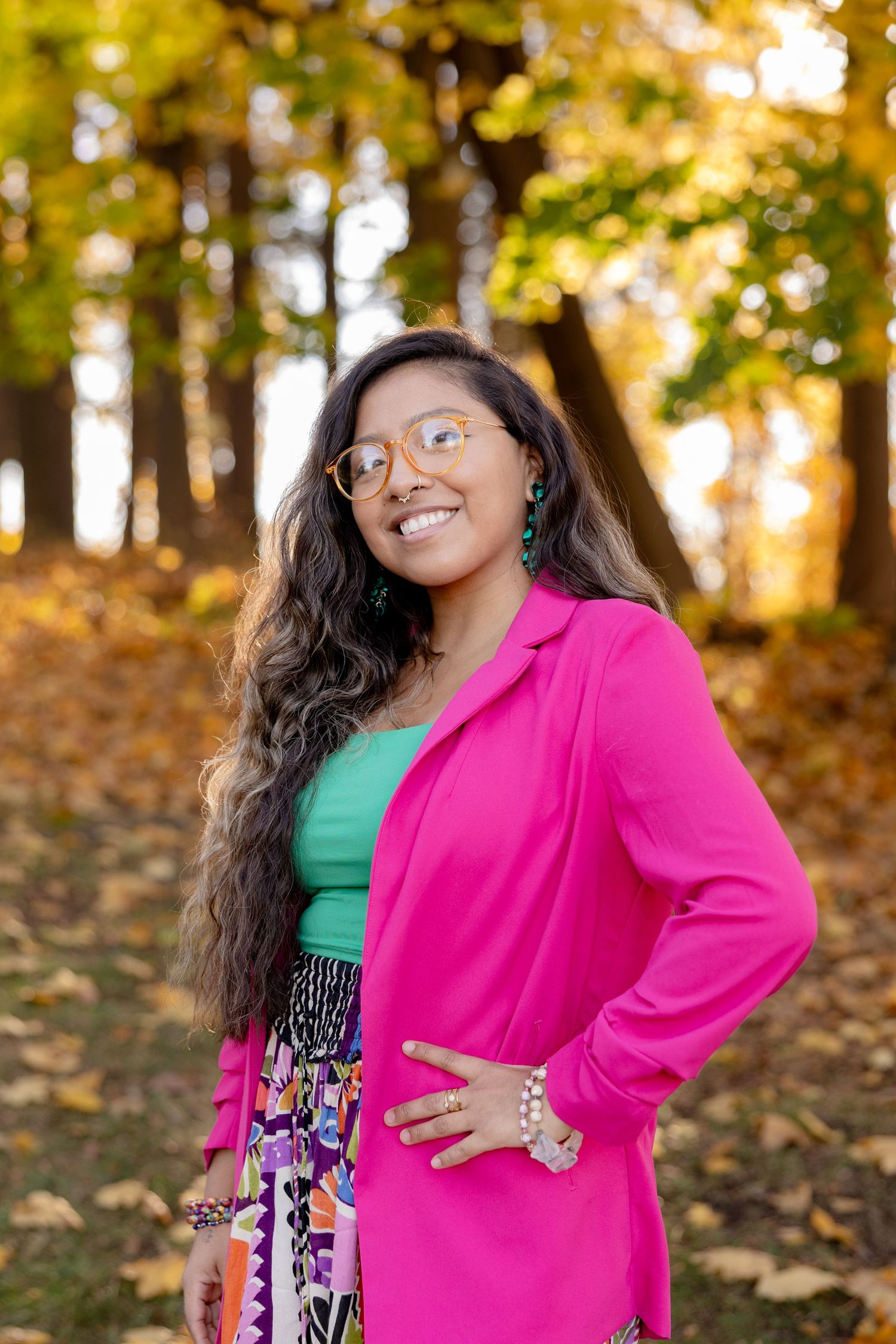 A person in a bright pink blazer and green top poses joyfully among autumn leaves in an outdoor setting.