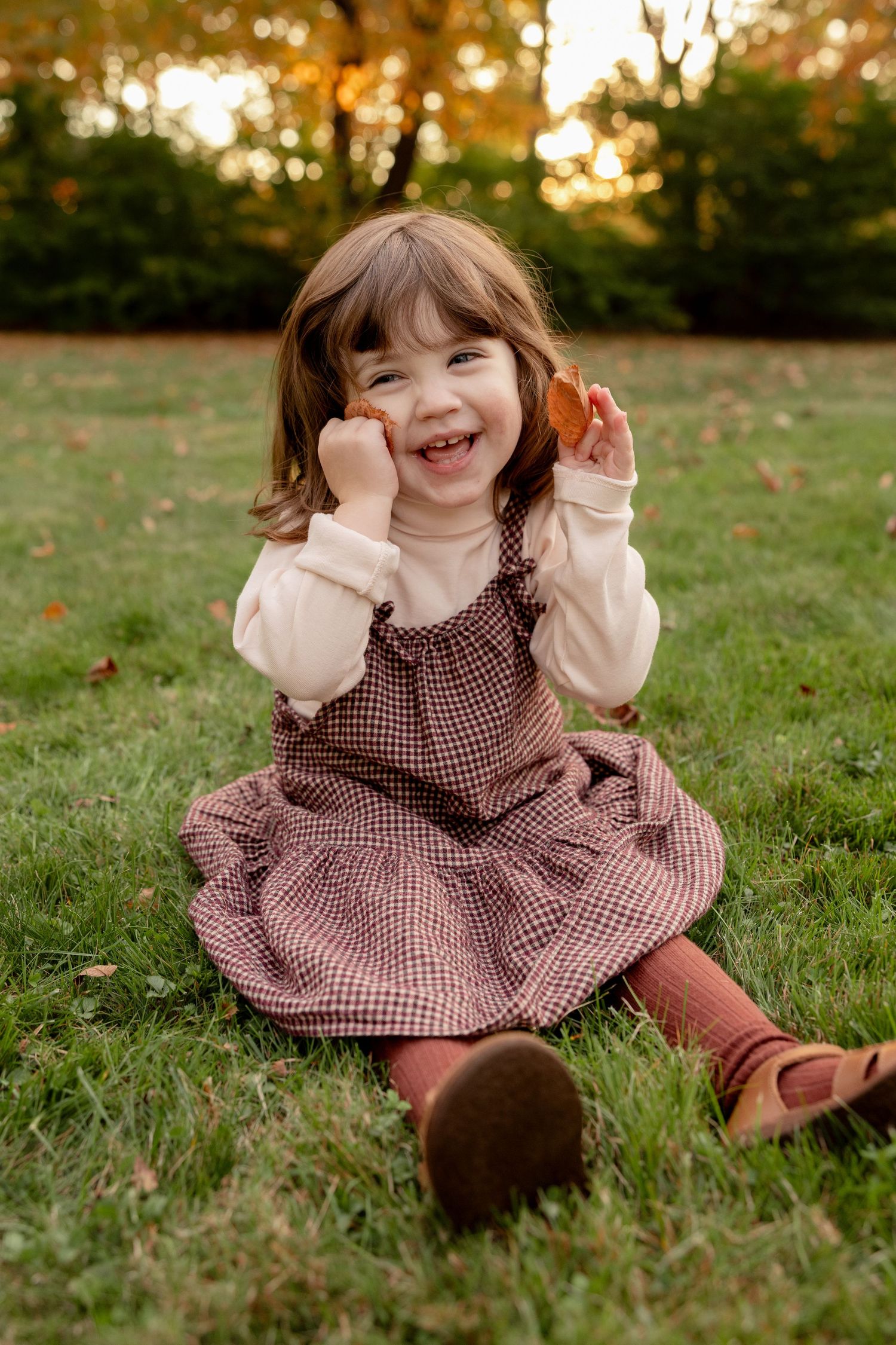 A child in a brown gingham dress sits on grass during golden hour, expressing pure joy and playfulness.