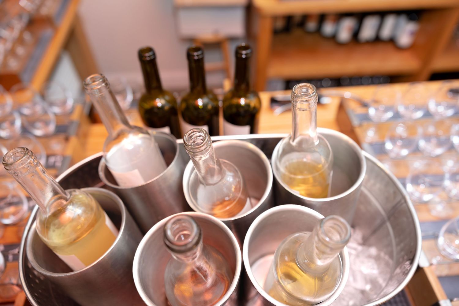 Wine bottles chilling in metal ice buckets with empty wine glasses arranged on a wooden surface.
