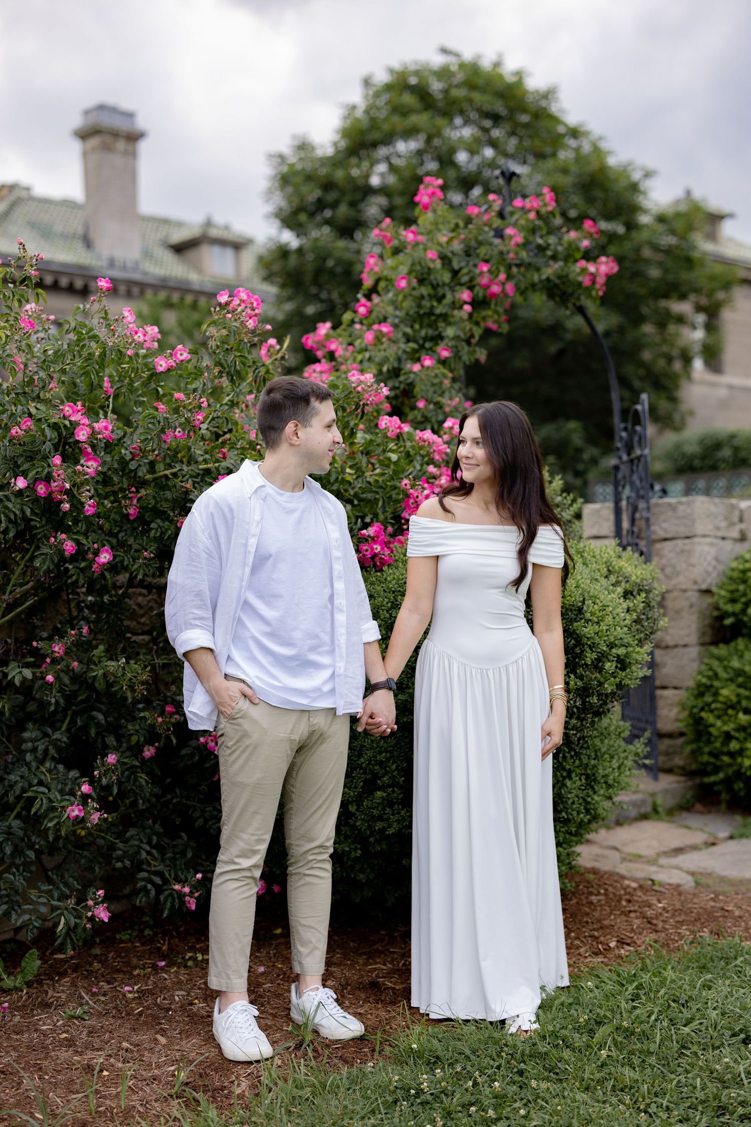 A romantic garden photoshoot featuring a couple in white outfits posing among blooming pink rose bushes.