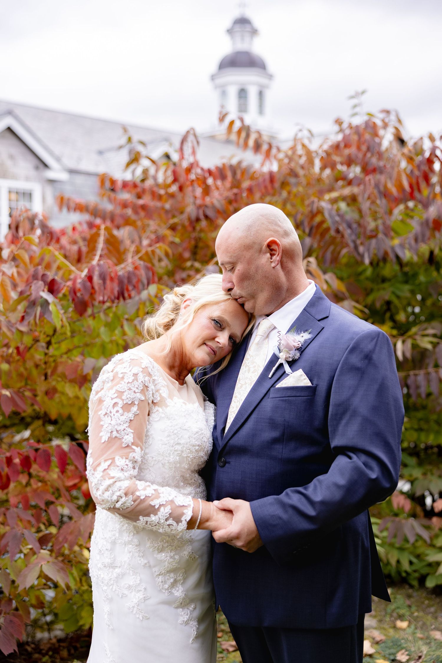A romantic autumn wedding scene with a couple embracing in front of red foliage and a white church steeple.