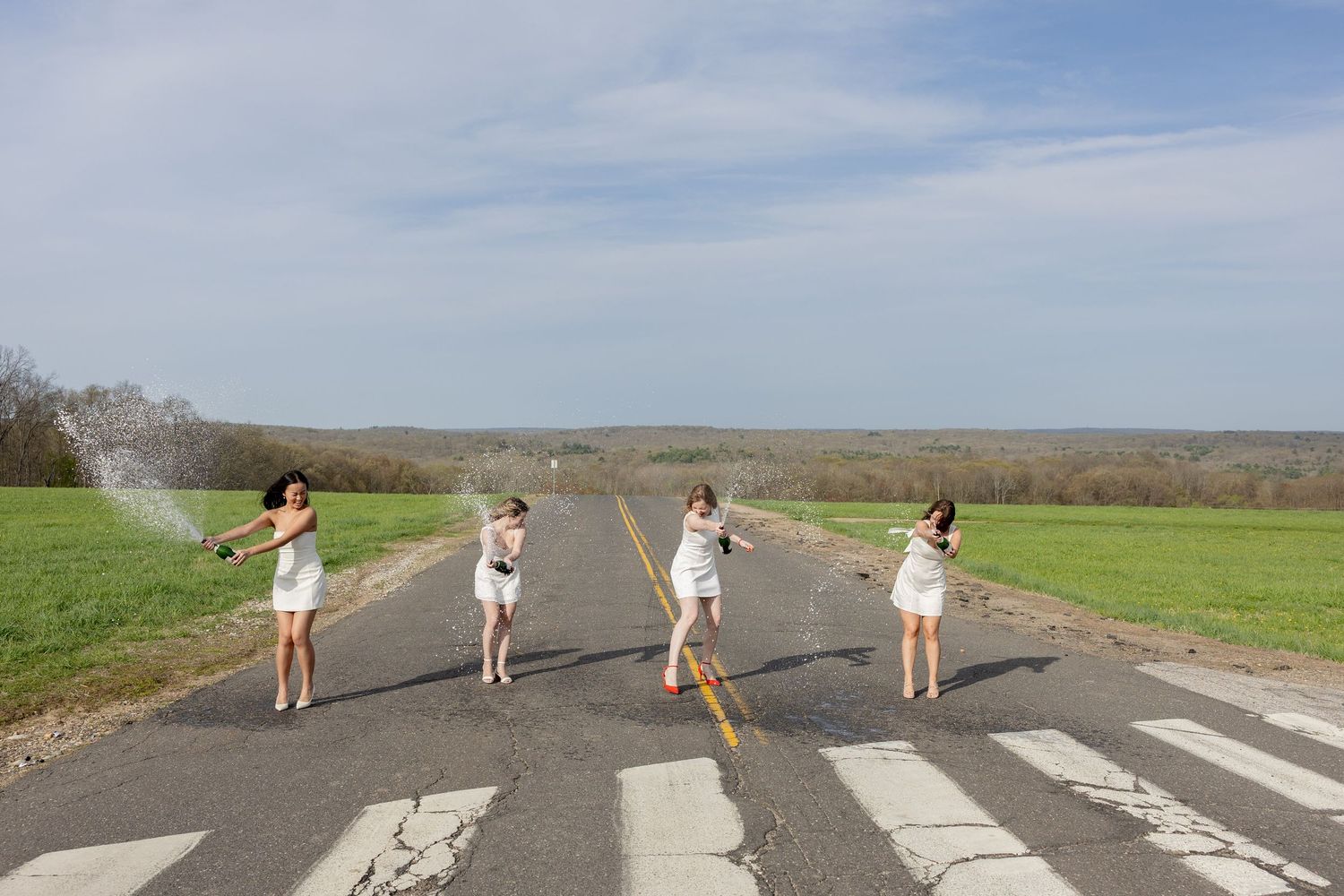 Four people pose on a crosswalk in summer outfits on a country road with green fields and blue sky in the background.