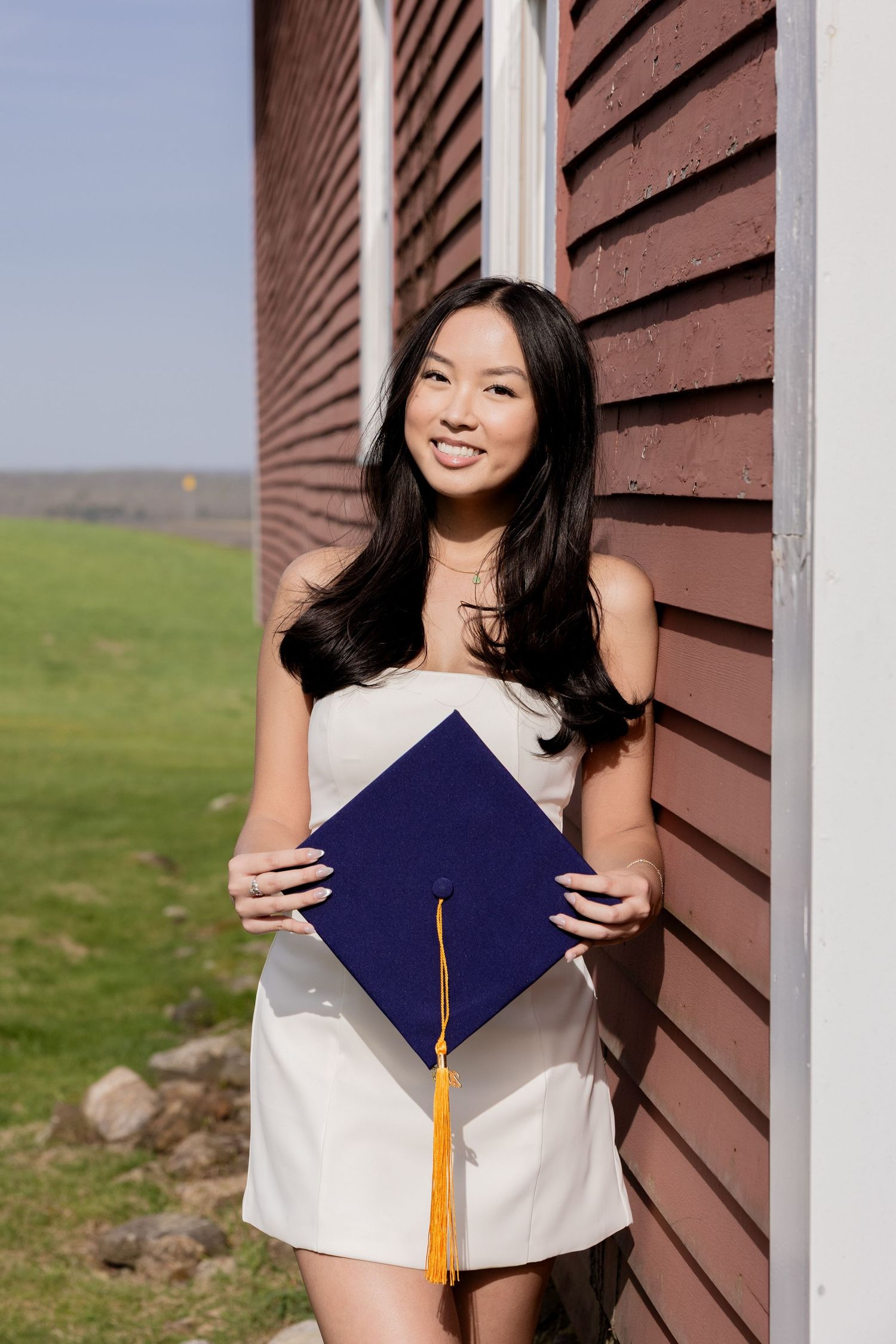Graduate wearing white dress holds blue graduation cap with gold tassel while standing next to rustic brown wall outdoors.