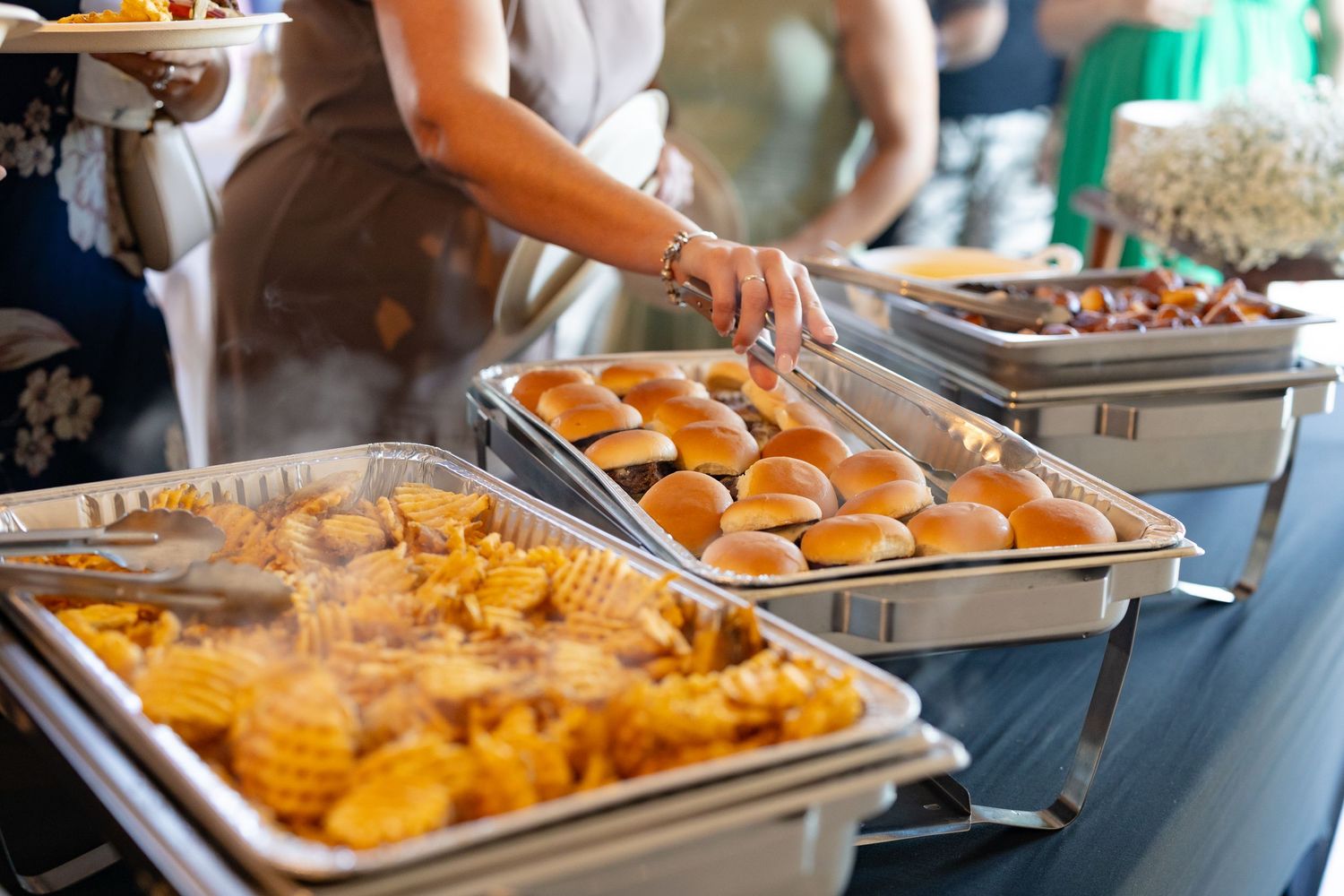 Buffet line of hot food being served in metal chafing dishes at catering event.