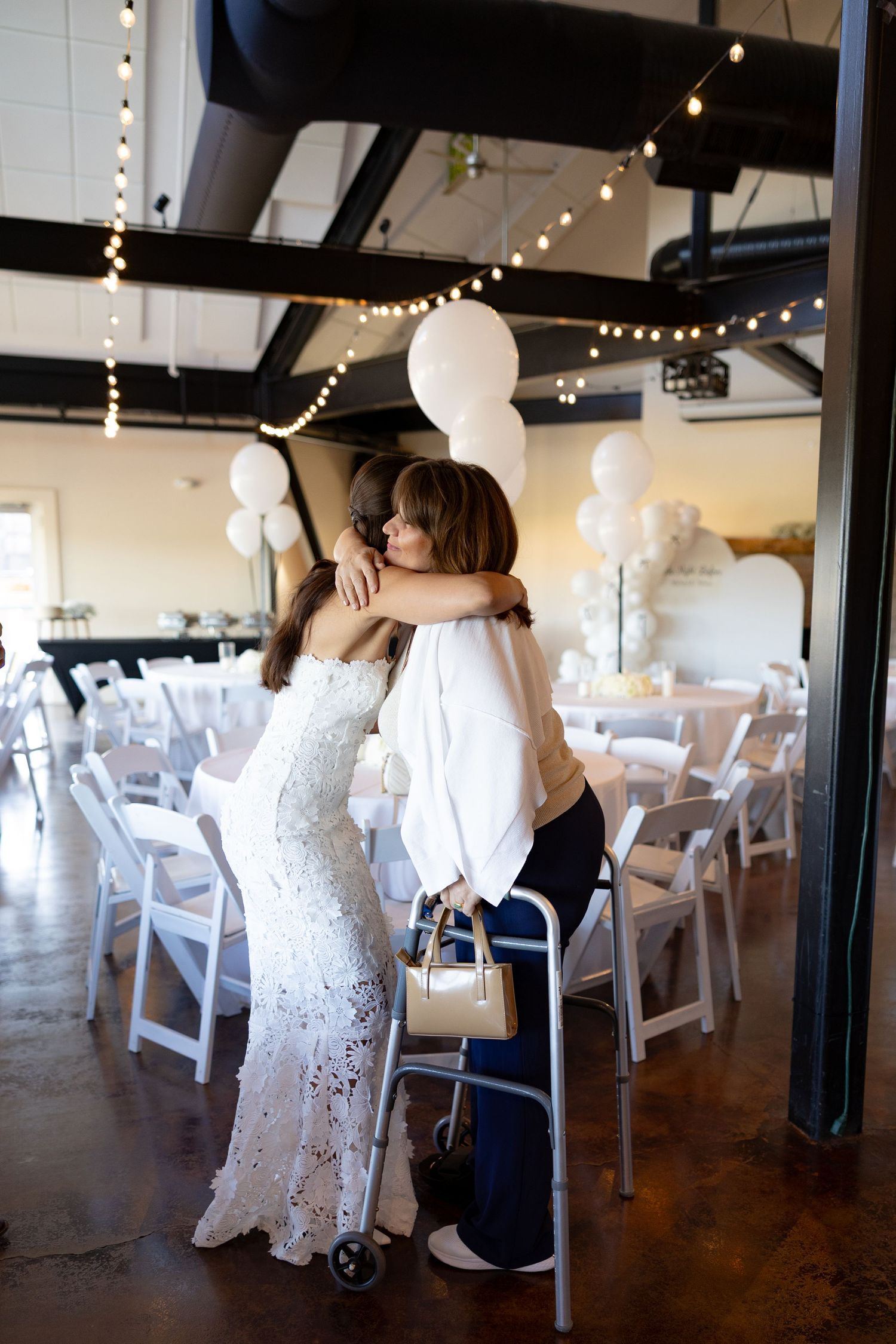 A touching moment between two people embracing in a wedding venue decorated with white chairs and hanging lights.