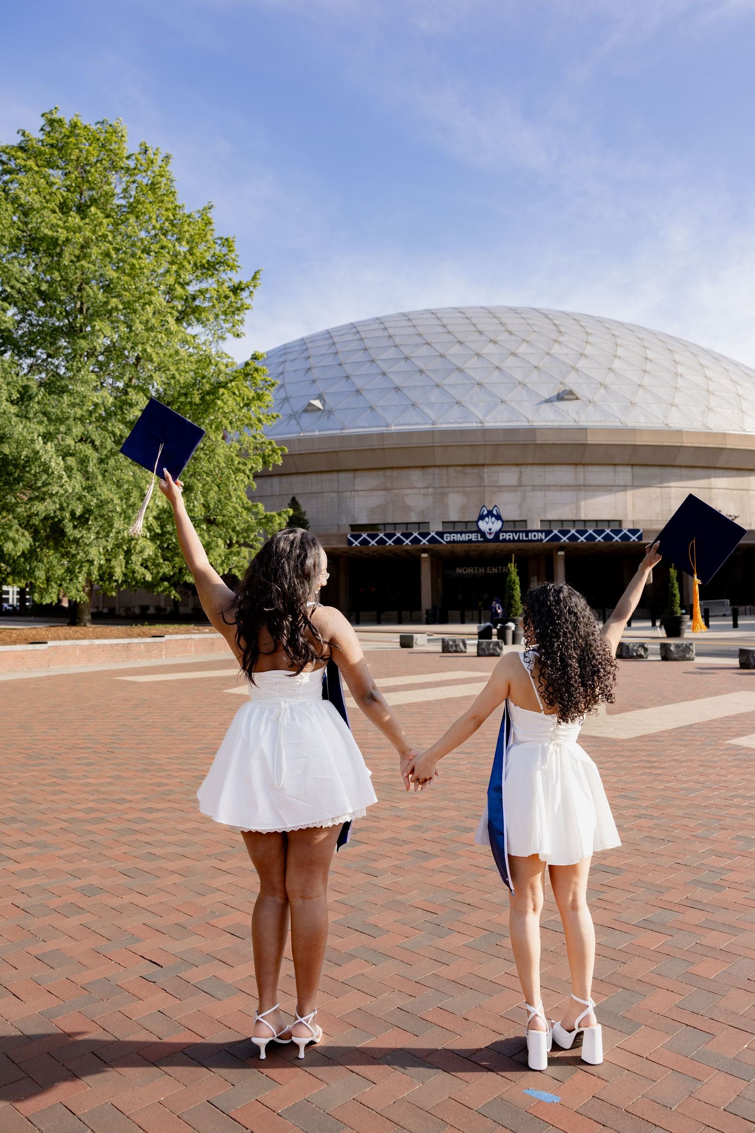 Two graduates in white and blue gowns celebrate their achievement outside a domed campus building on a sunny day.