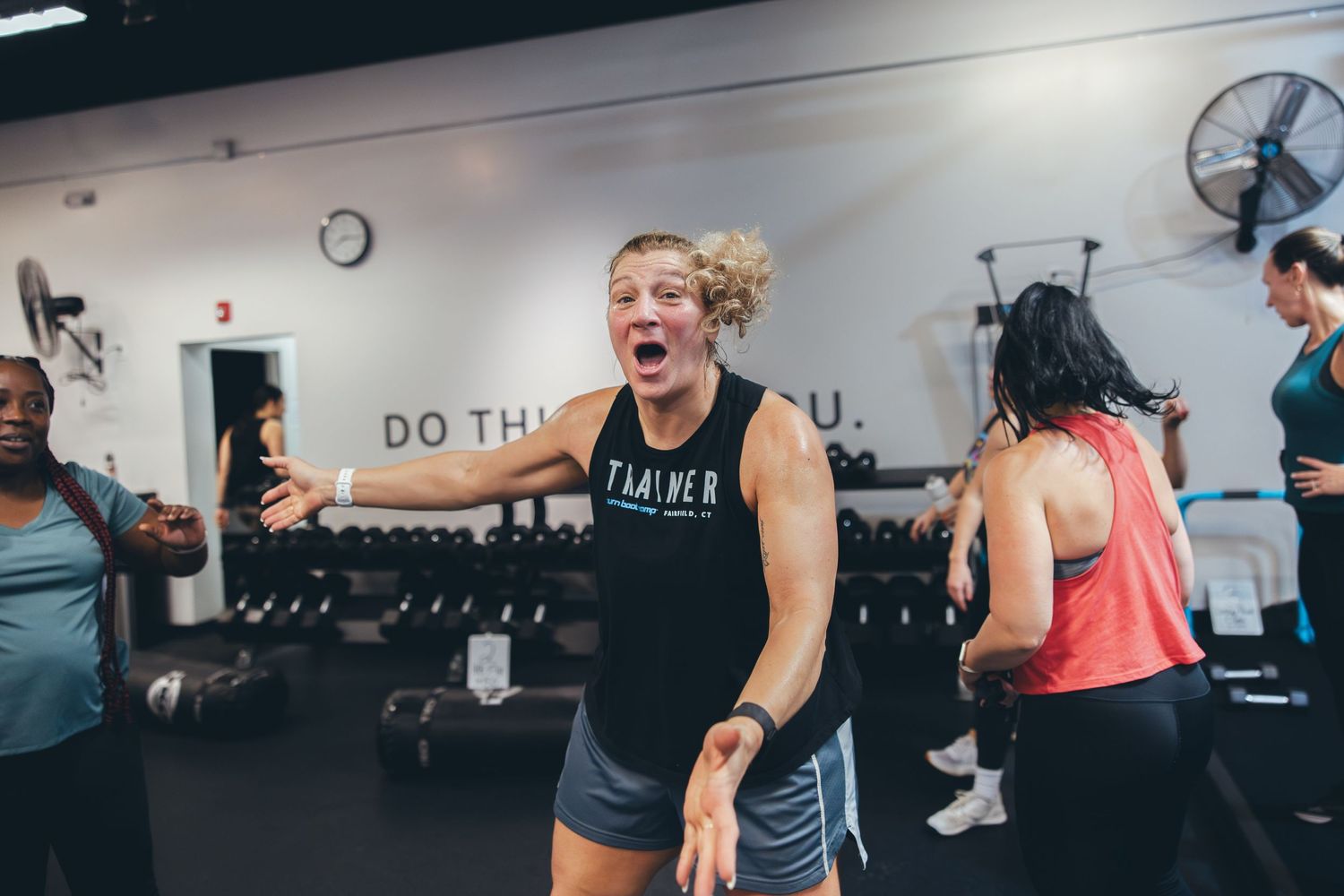 People exercising in a gym with a wall of dumbbells and motivational text visible in the background.