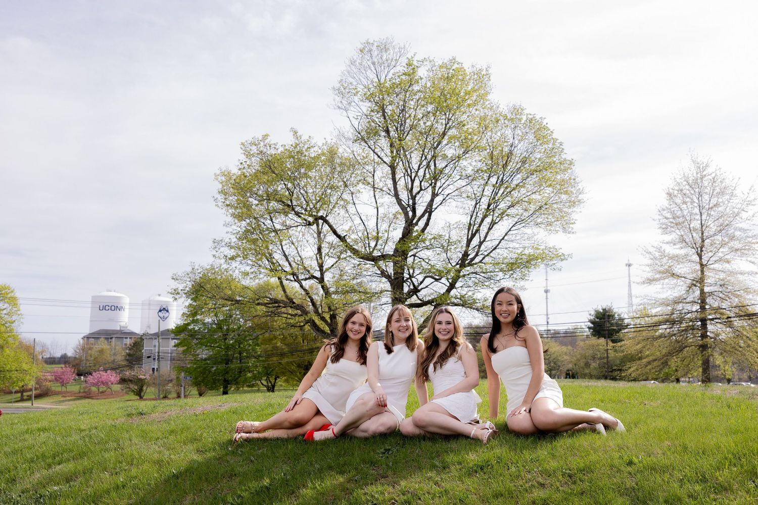 A group sits together on a grassy hill in spring, wearing white dresses with a large tree and city skyline in the background.