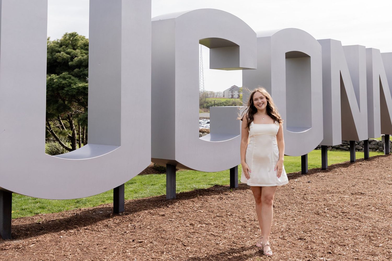 A young person in a white dress stands next to large white UCONN letters outdoors on a path.