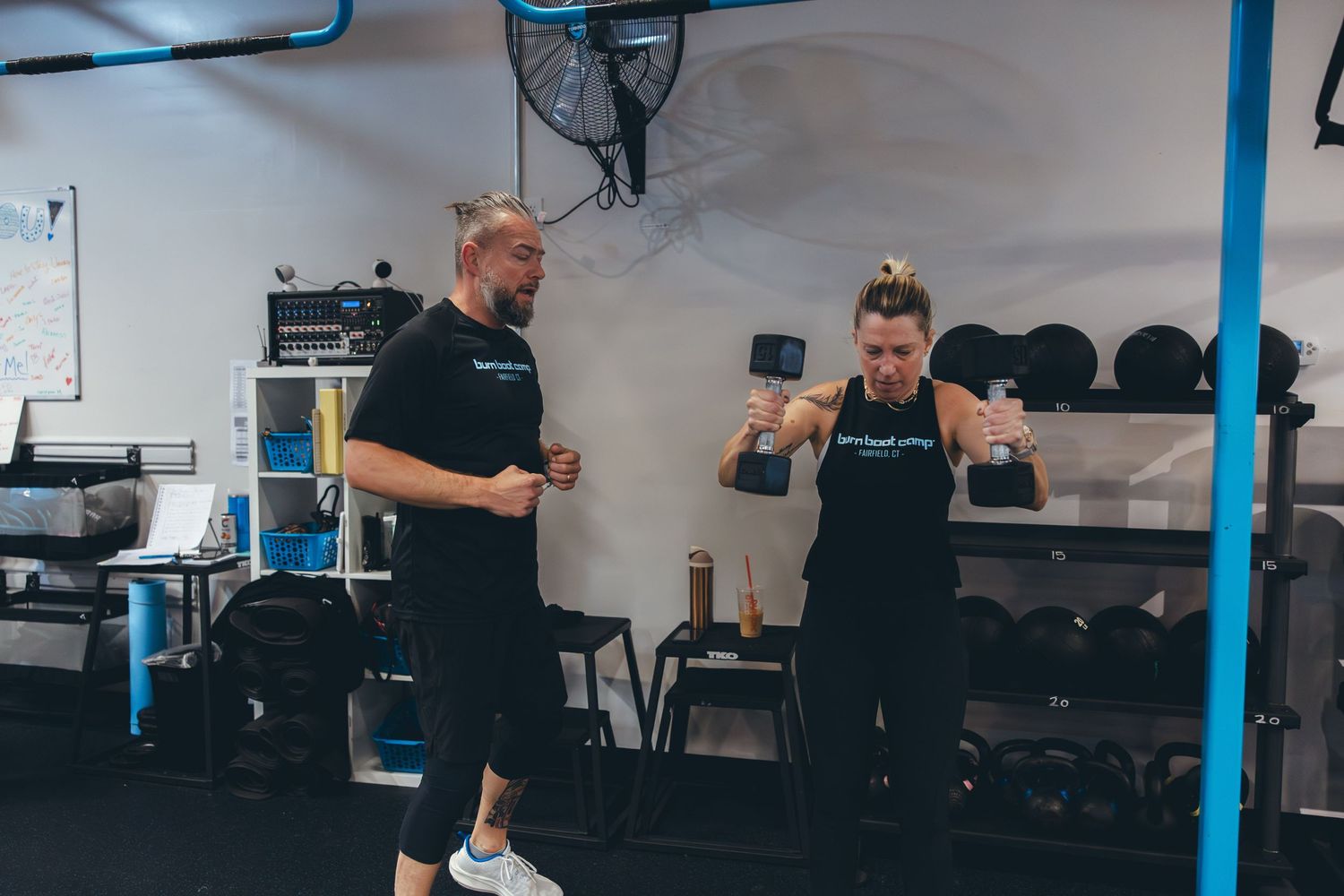 Two fitness trainers demonstrating exercises in a gym with equipment storage area visible.