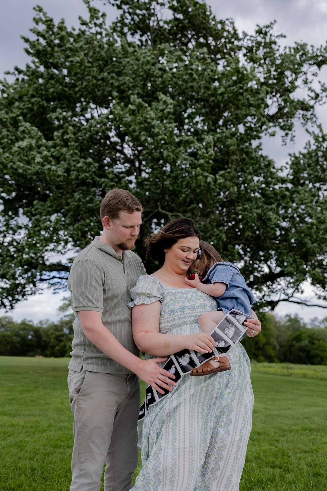 A tender family moment outdoors under a leafy tree with a small child and expecting parents.