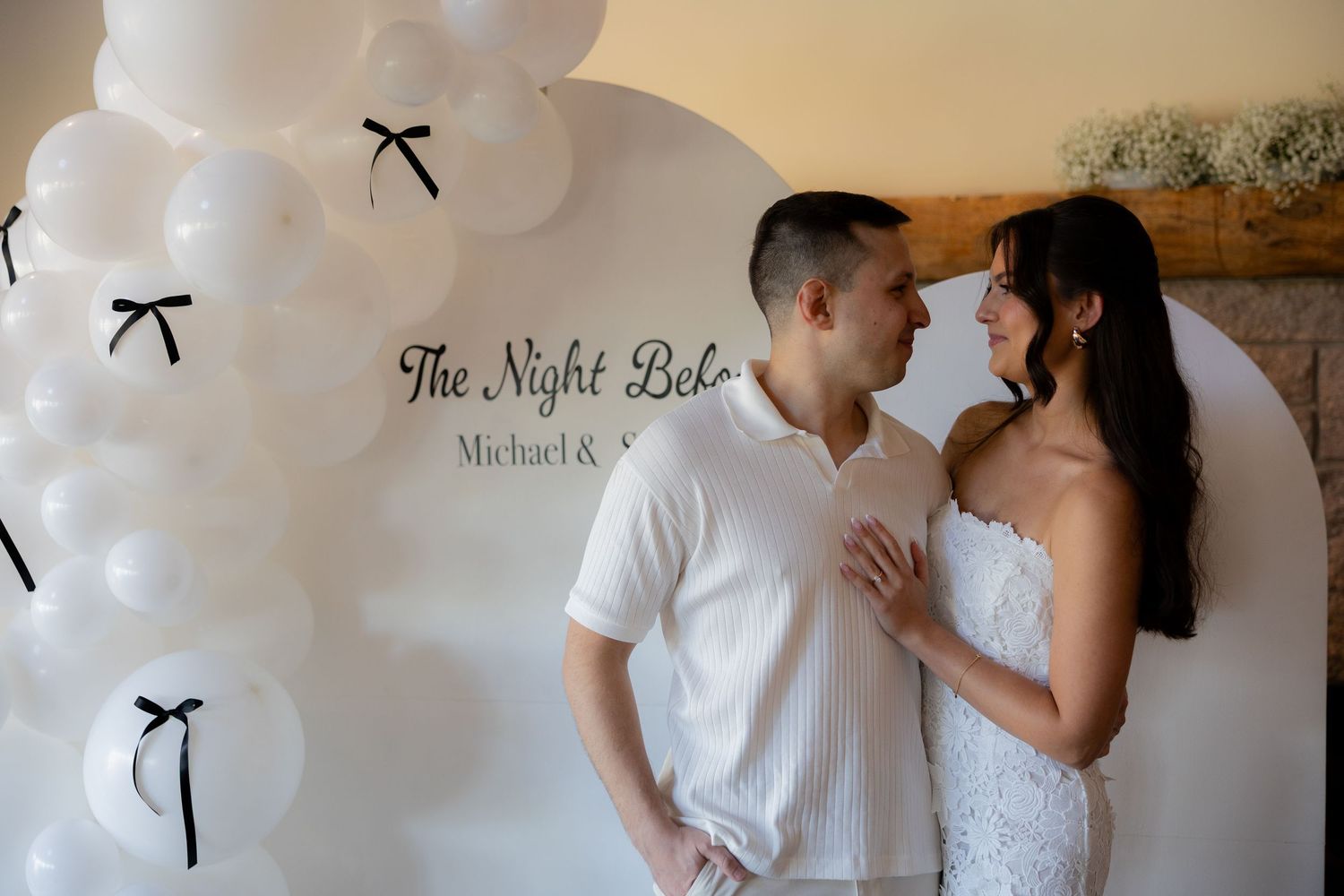A wedding couple stands near white balloons with black ribbons at an event marked 'The Night Before'.