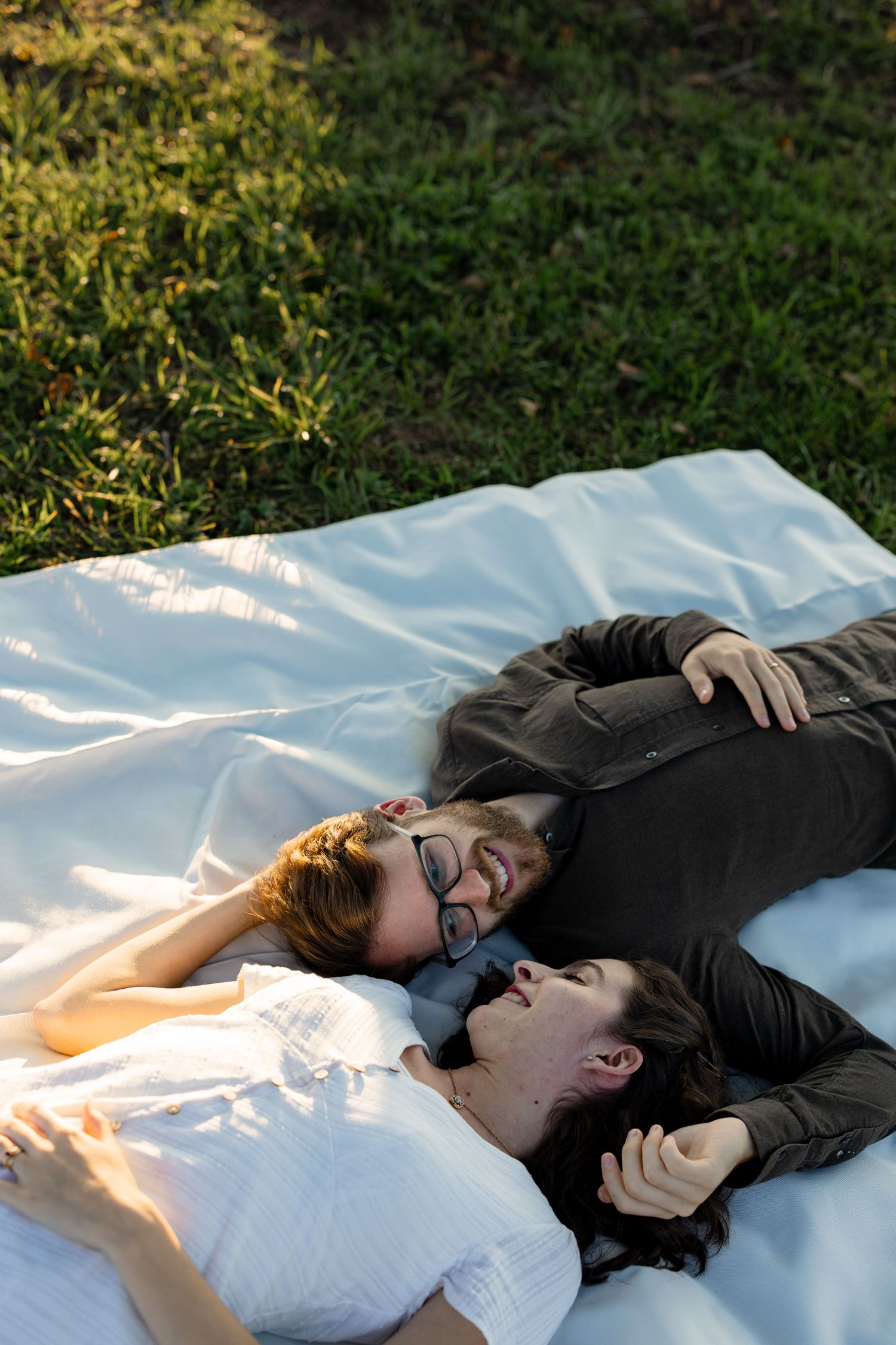 A couple lies together on a white blanket outdoors in the grass during golden hour sunlight.