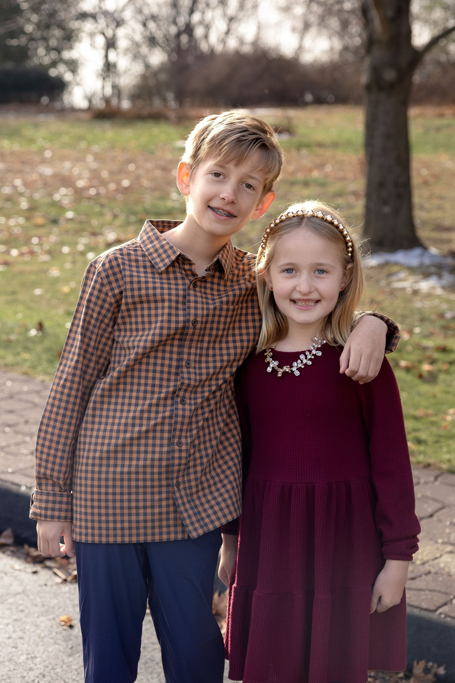 Two children in coordinating outfits of burgundy dress and brown plaid shirt pose together outdoors in autumn.