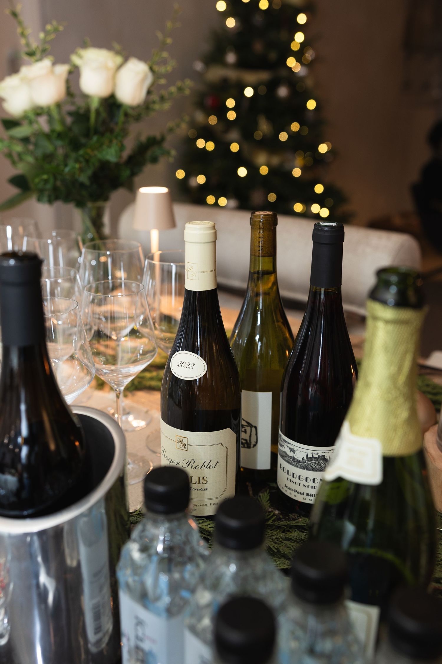 Wine bottles and glasses arranged on a table with twinkling holiday lights in the background.