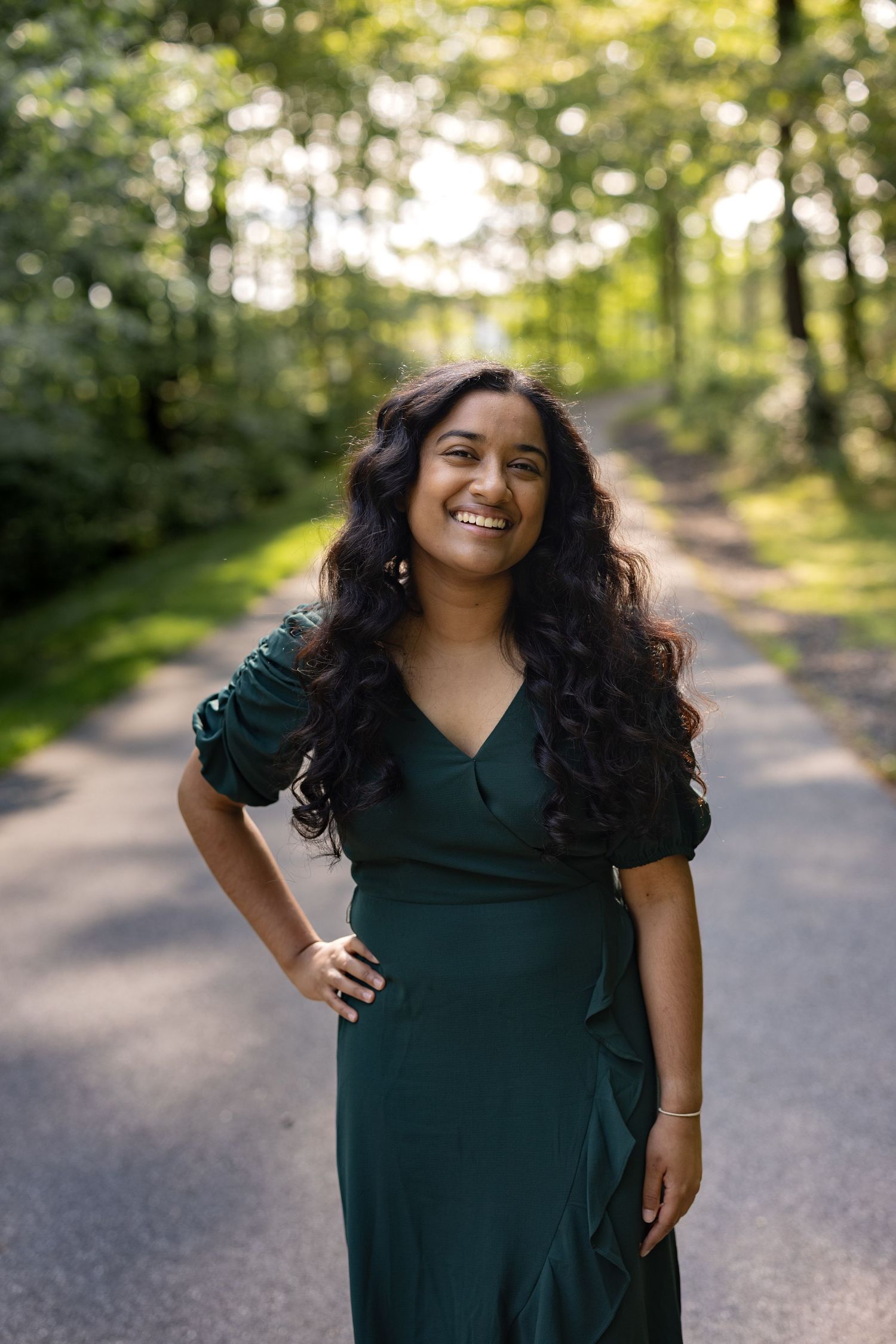 A cheerful individual in a dark green wrap dress poses on a sunlit forest path with flowing dark hair.