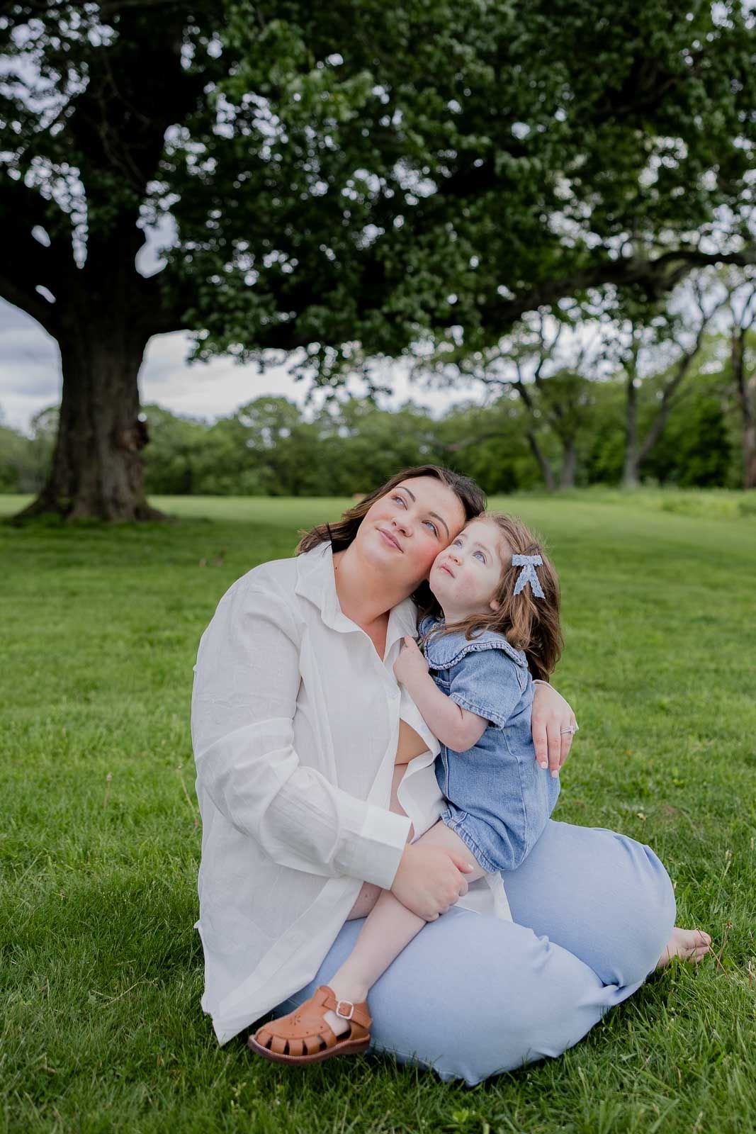 Parent and child sitting together on green grass under a large tree in a peaceful park setting.