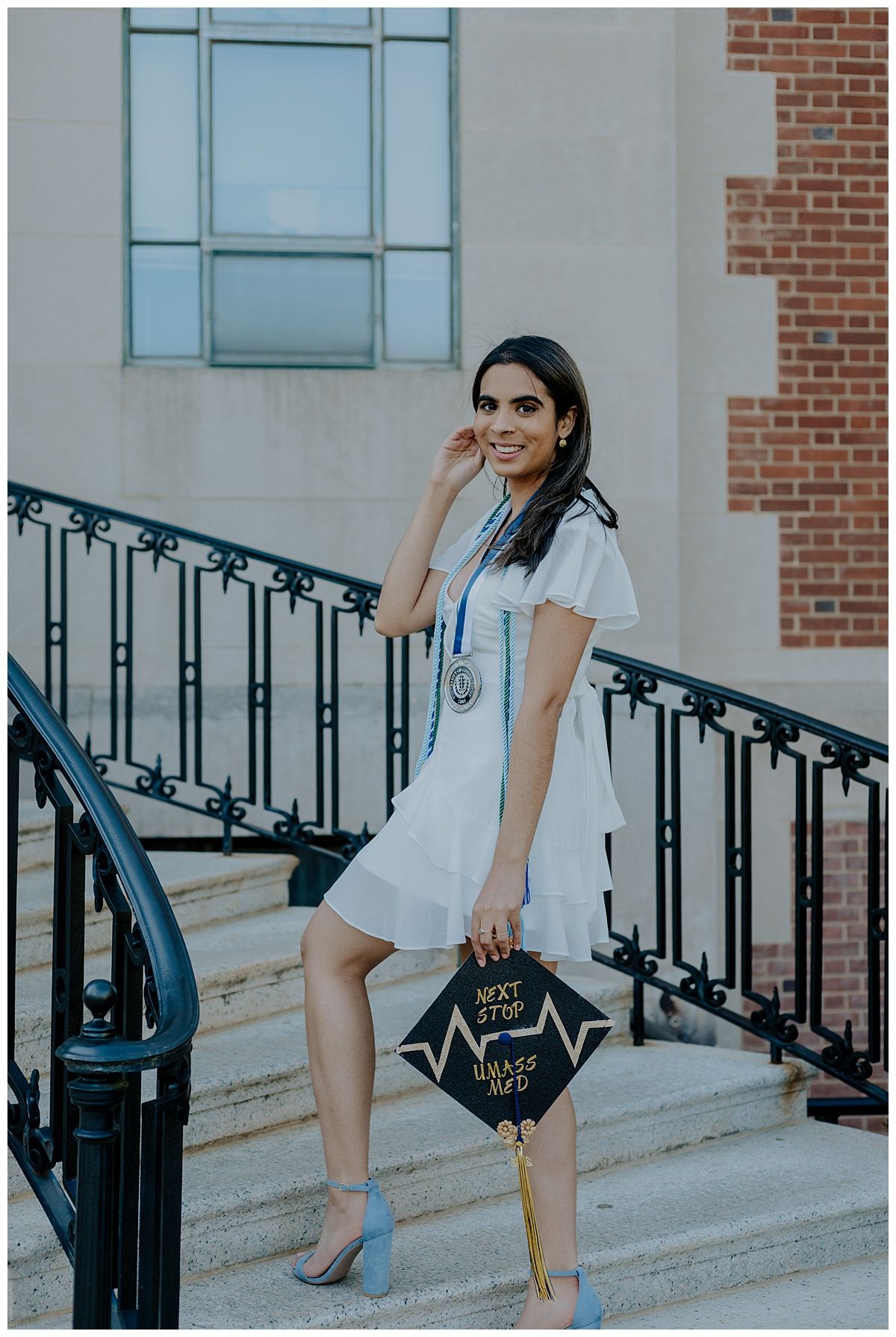 Graduate in white dress poses on stairs with decorated cap during commencement photoshoot.