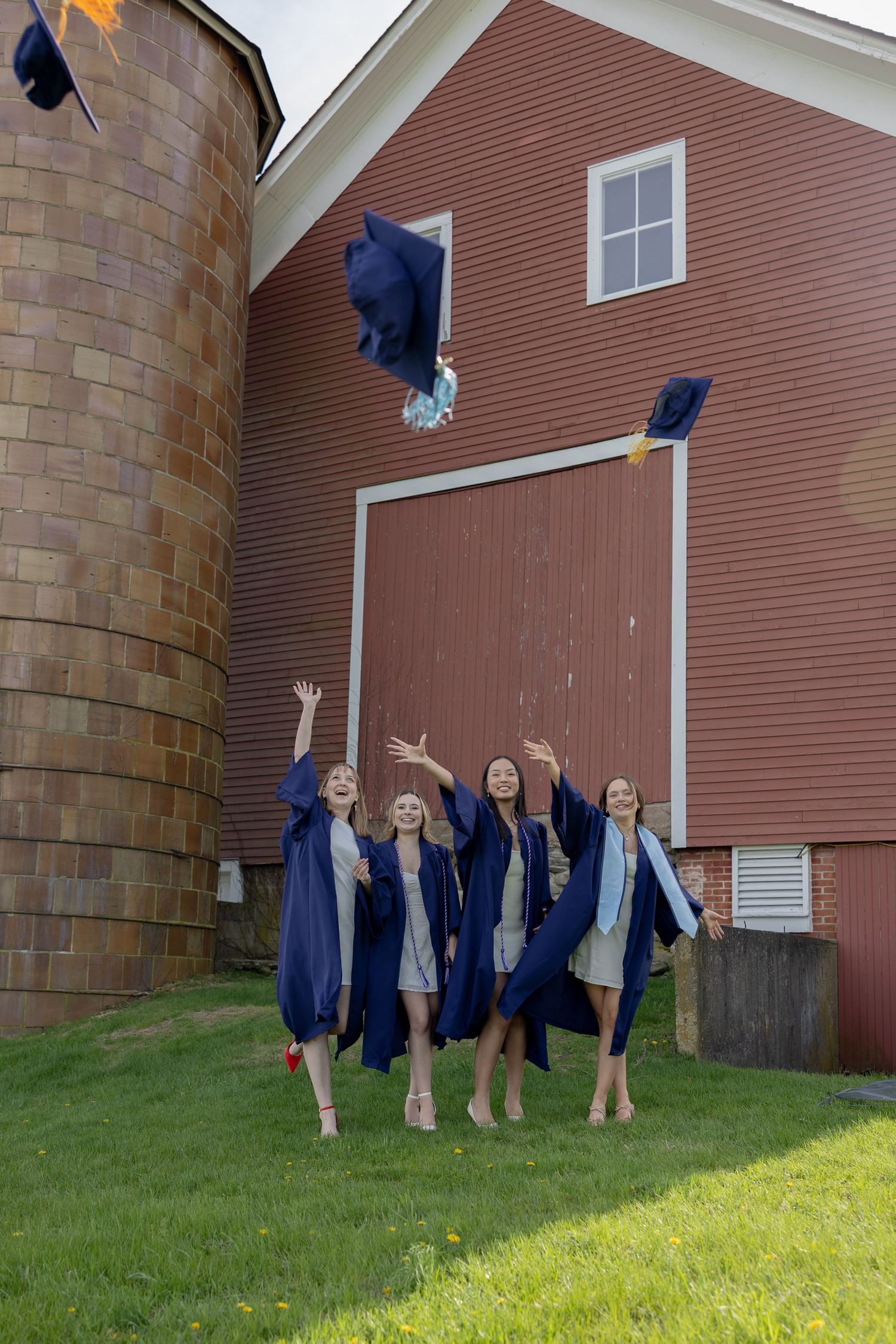 Graduates in blue caps and gowns throw their mortarboards in celebration outside a rustic red barn with silo.