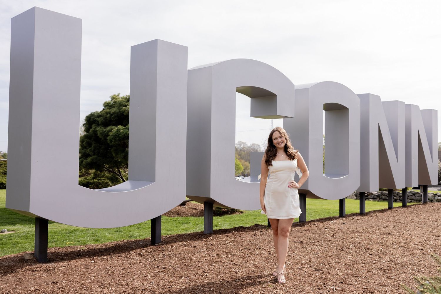 Someone in a white dress poses beside giant white UCONN letters on a dirt path with greenery in the background.