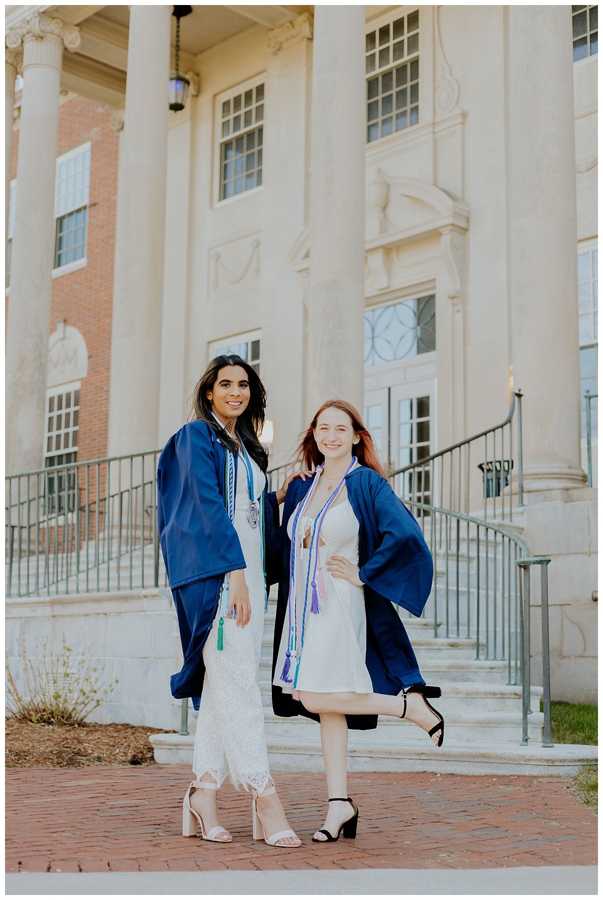 Two graduates in blue caps and gowns pose on steps of classical building.