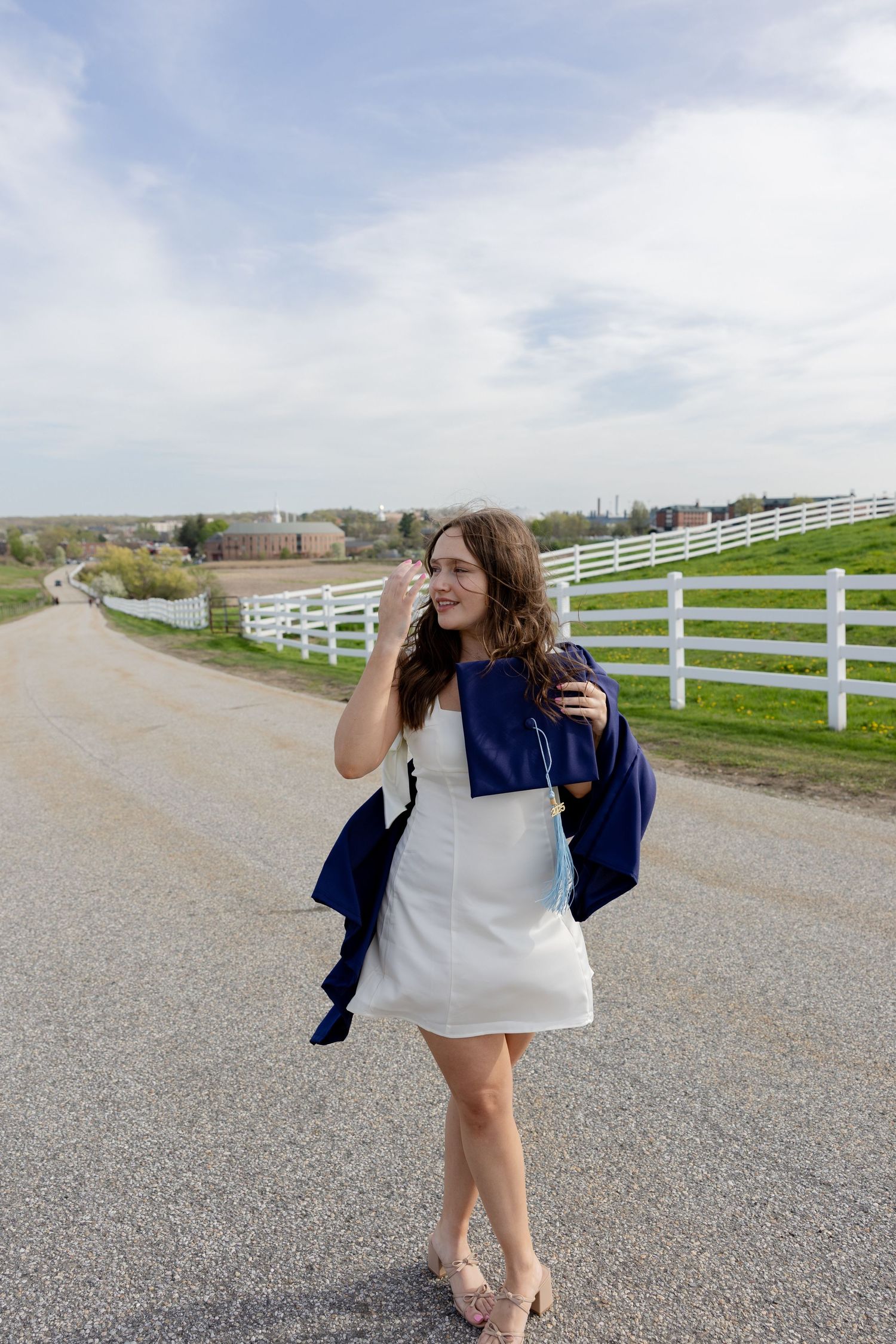 A person wearing a white dress and navy cardigan stands on a country road with white fences in the background.