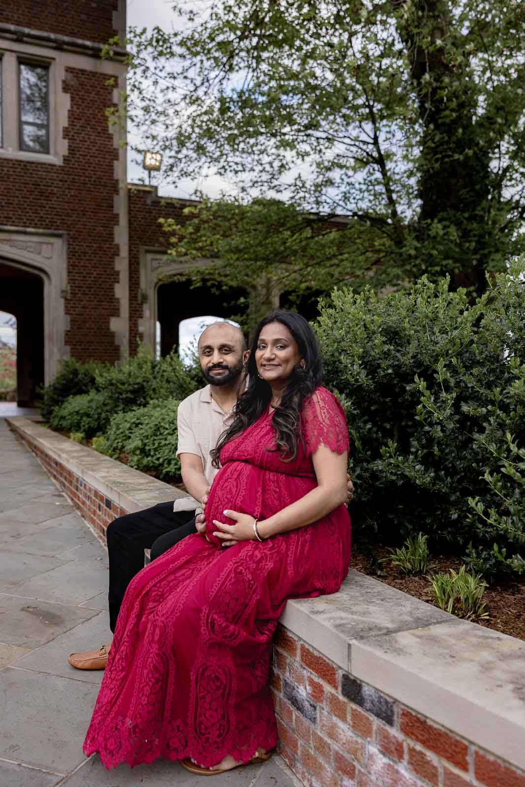 Couple sits on brick ledge by building with woman in red maternity dress holding her baby bump.
