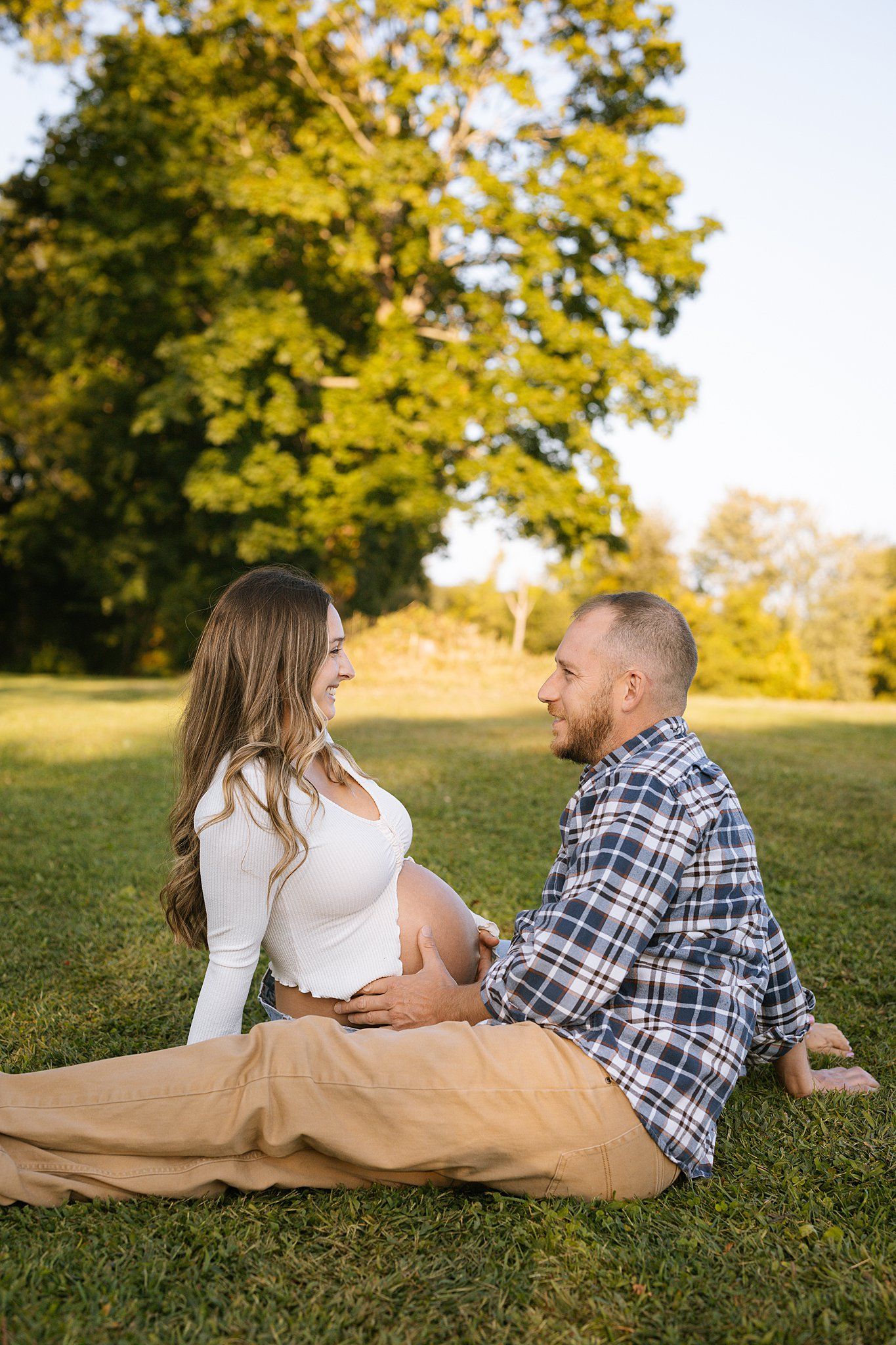 Expecting couple shares intimate moment on blanket in sunny park setting.
