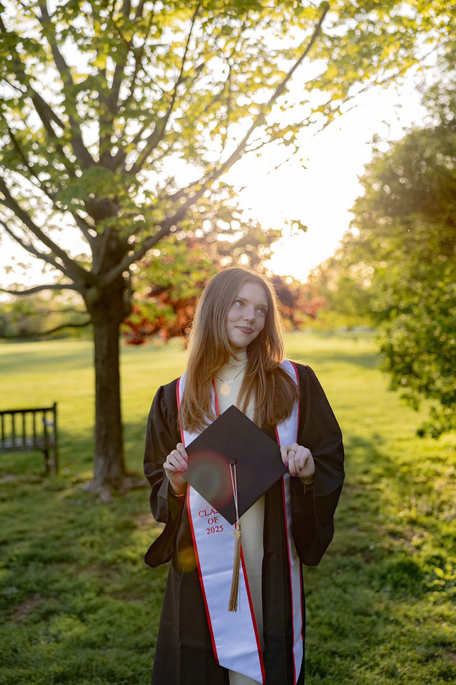 Graduate holds diploma while standing in a grassy field at sunset with trees in the background.
