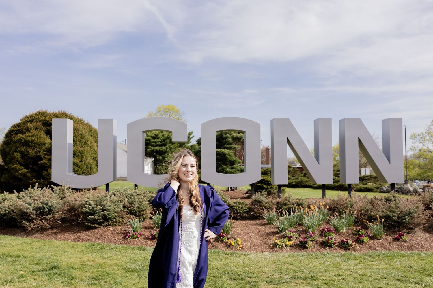 Someone poses in front of large white UCONN letters on a grassy lawn with shrubs and trees in the background.