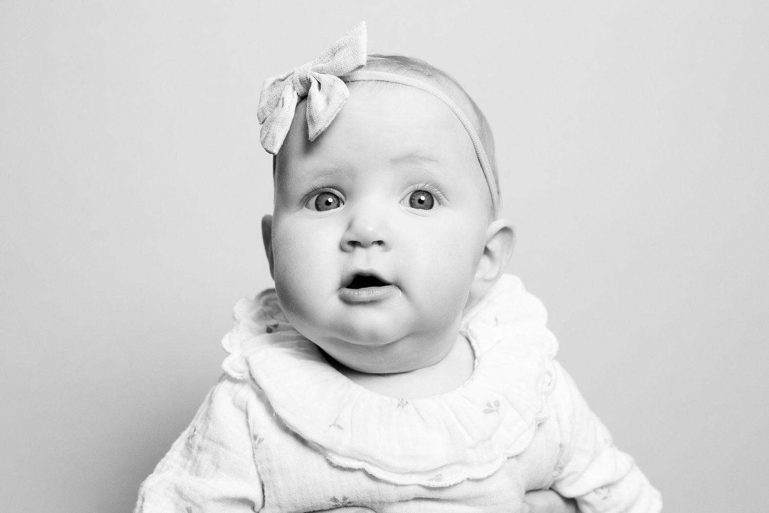 Black and white portrait of a baby wearing a white outfit and bow headband with an expressive look.
