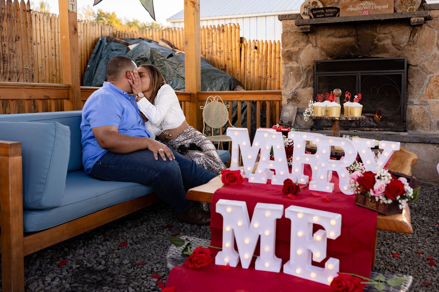 Red and white 'MARRY ME' letters arranged outdoors with romantic lighting and rustic decor for a proposal setup.