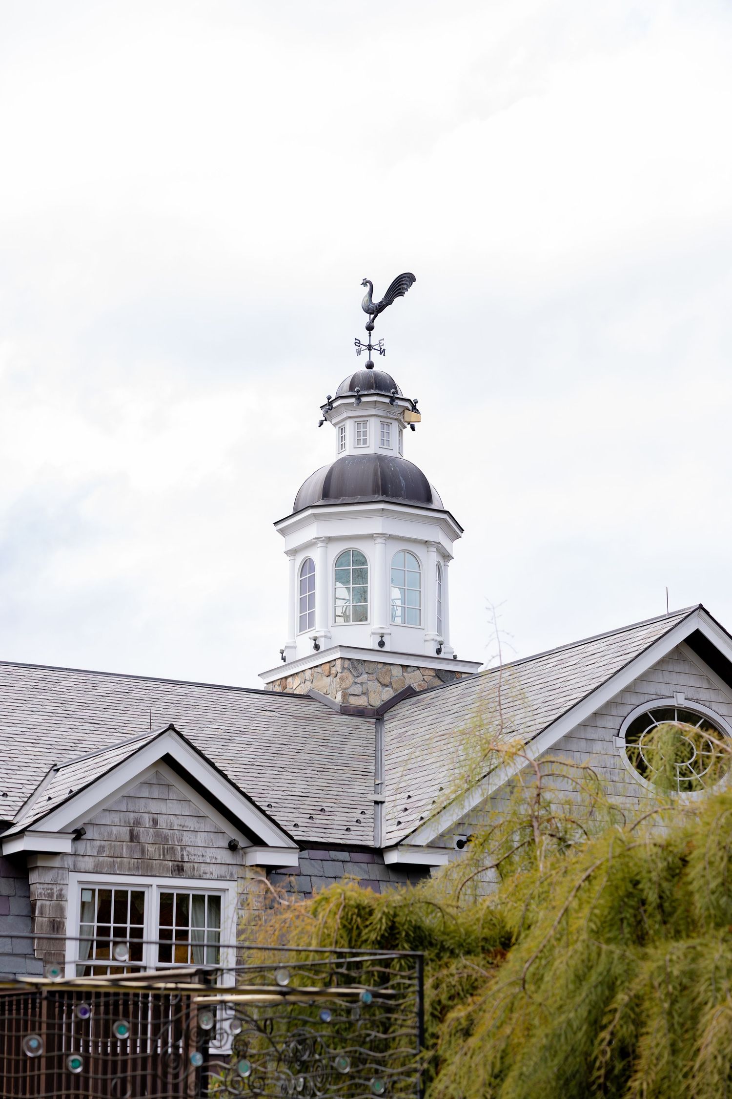 White cupola with weather vane sits atop gray shingled building with arched windows and willow trees in foreground.