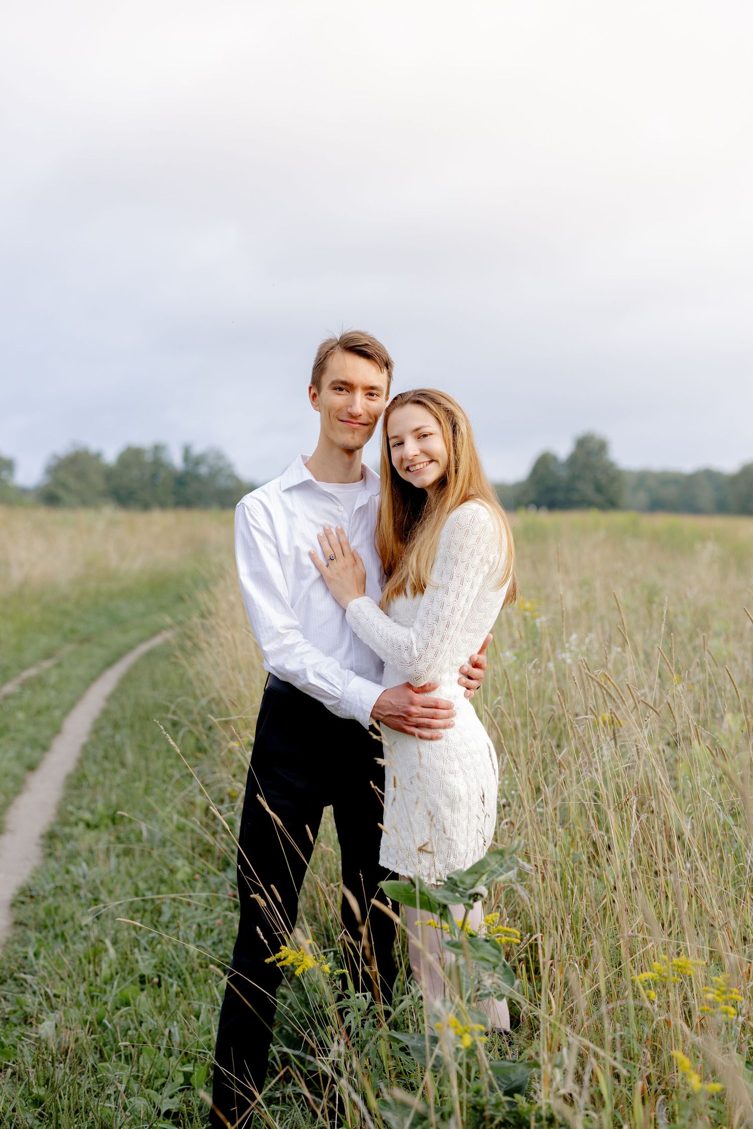 A couple embraces lovingly in a serene meadow with tall grass and wildflowers on a sunny afternoon.