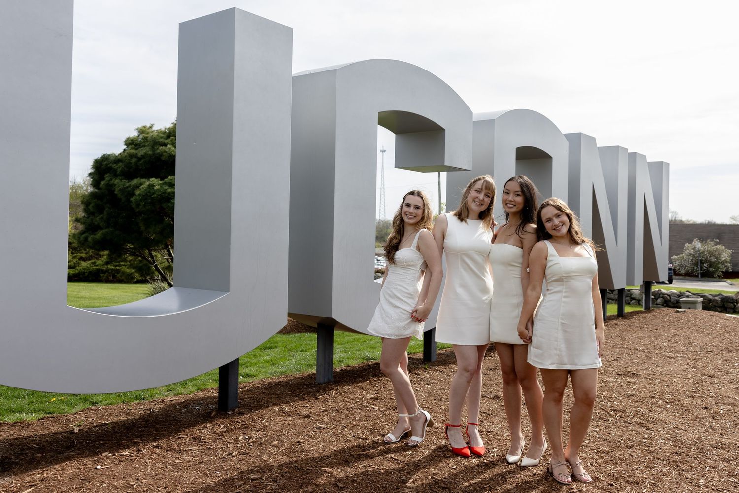 Three people in white dresses pose together in front of large white letters on a hillside.
