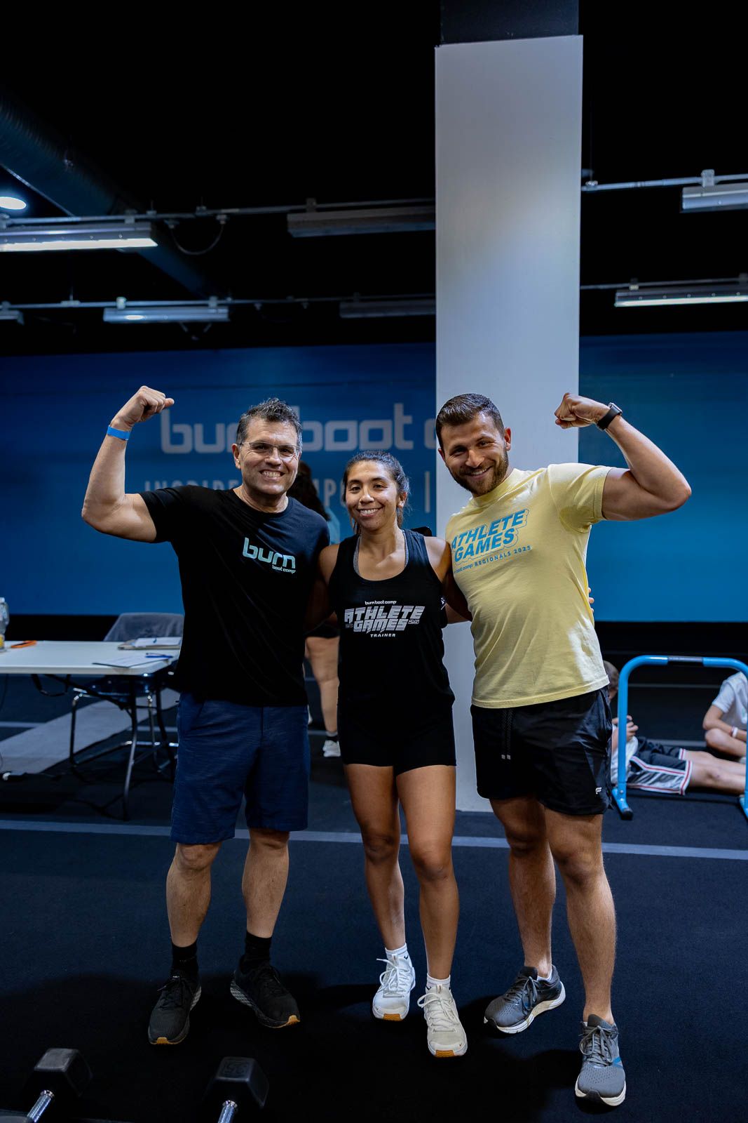 Three fitness enthusiasts flex their muscles and pose together at a gym with blue walls.