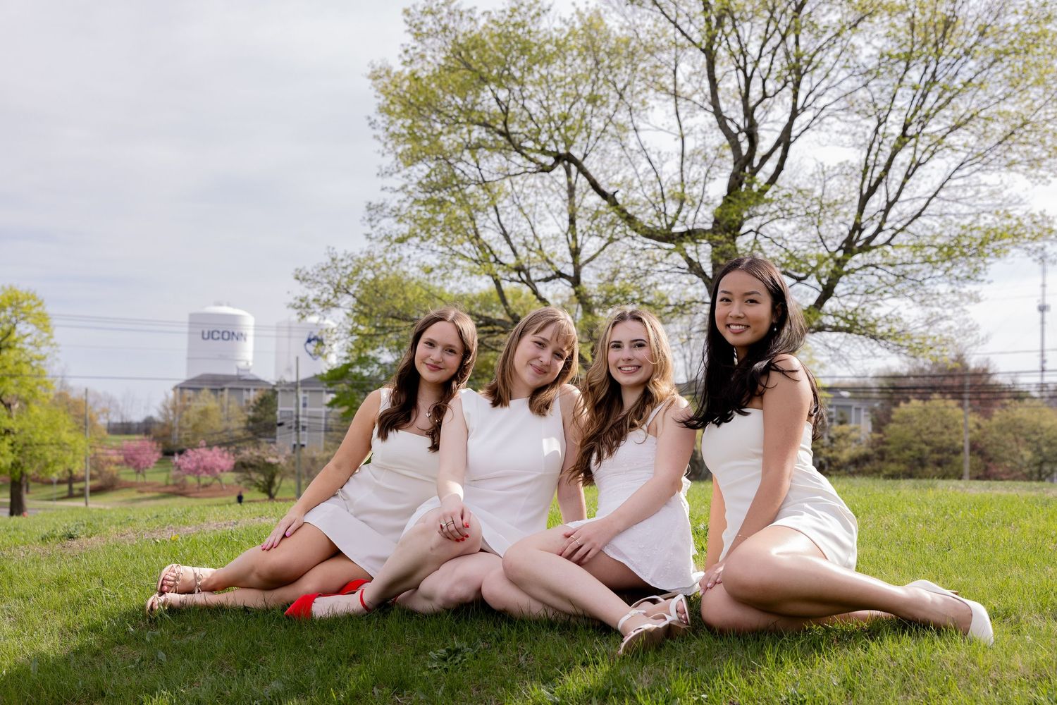 A group in matching white dresses sits together on grass with a city skyline visible in the background.