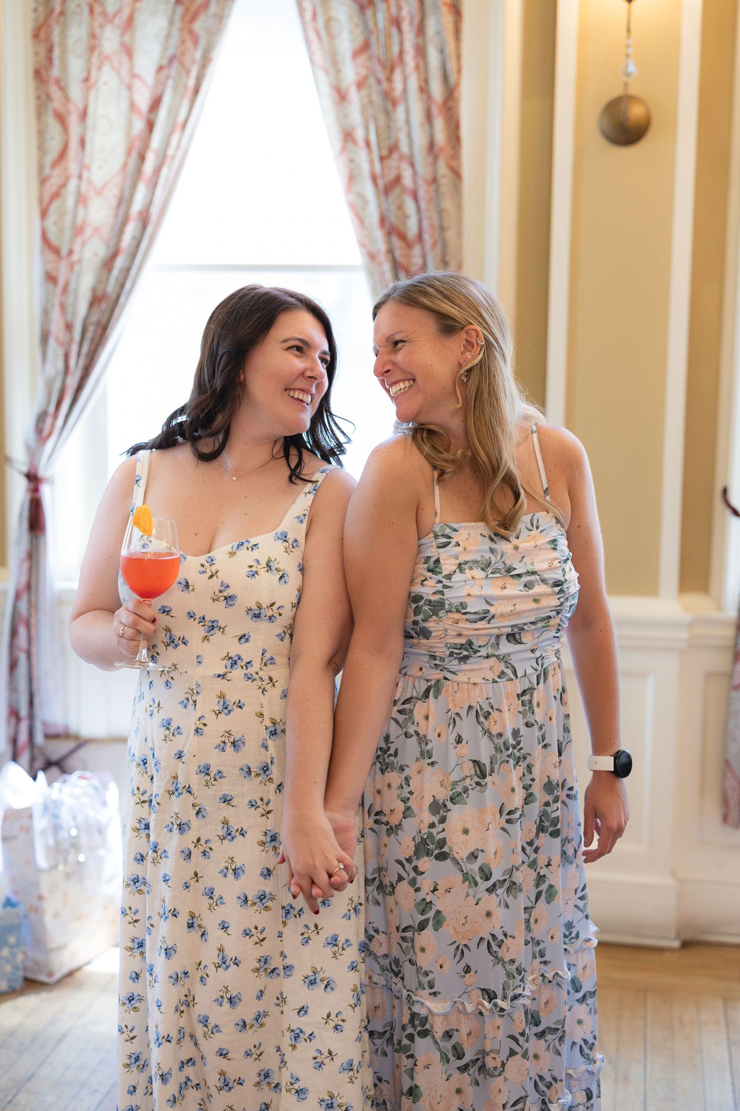 Two women in matching floral dresses laugh and hold hands while standing in an elegant room with floral curtains.