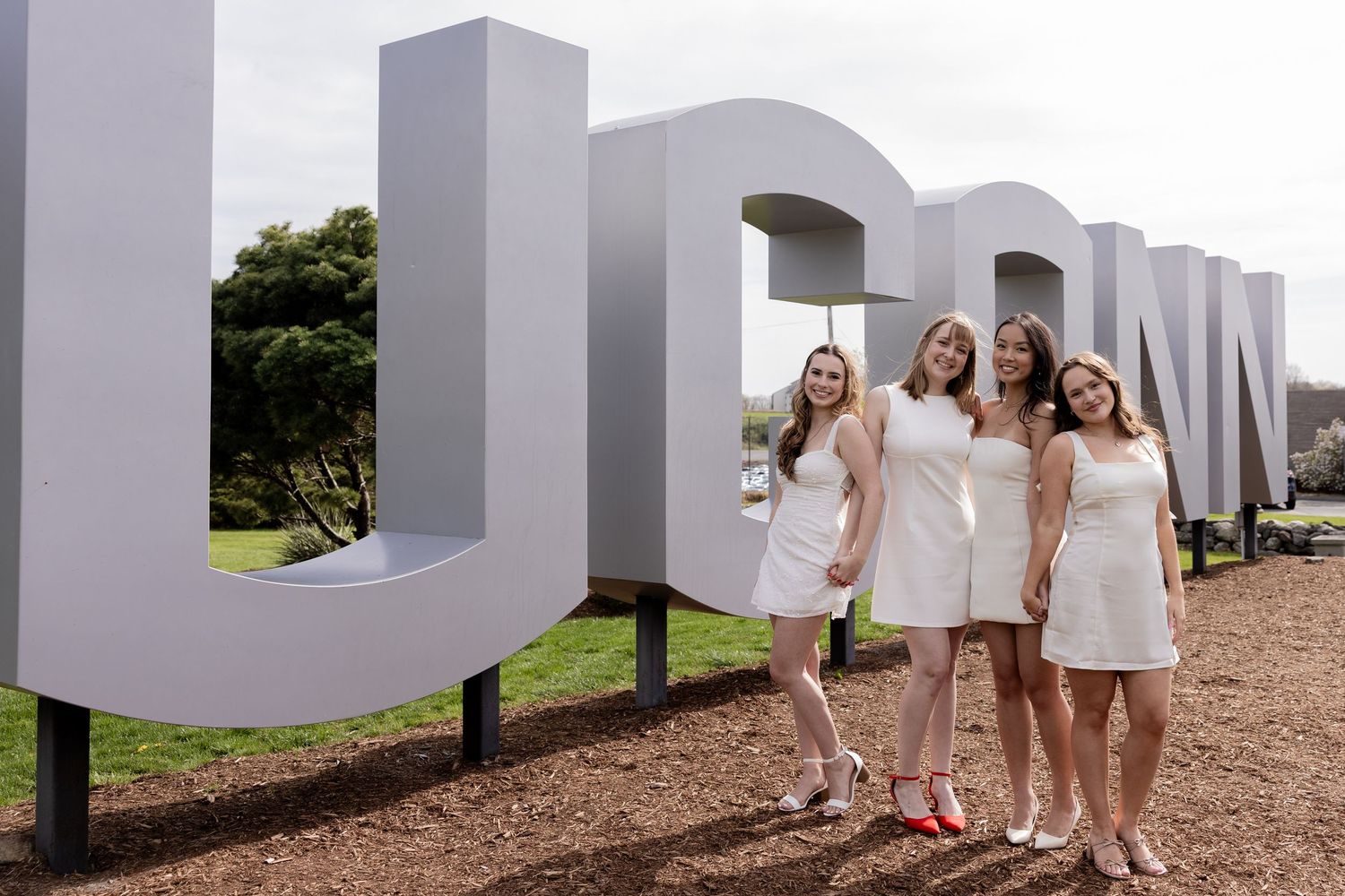 Four people in white dresses pose together in front of large white UCSD letters outdoors on a cloudy day.