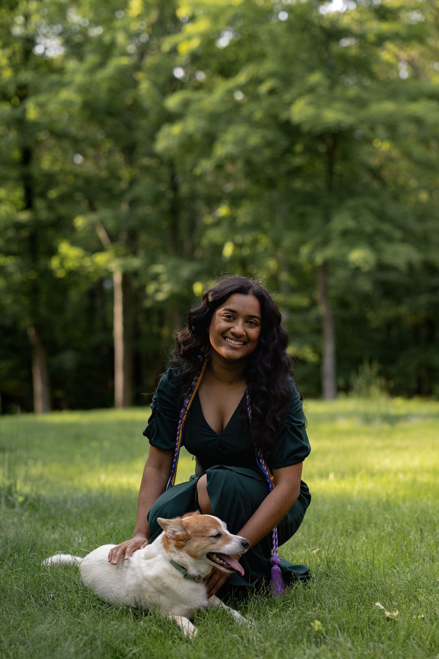 A joyful woman in a green dress sits in a grassy park while petting a white dog laying beside her.